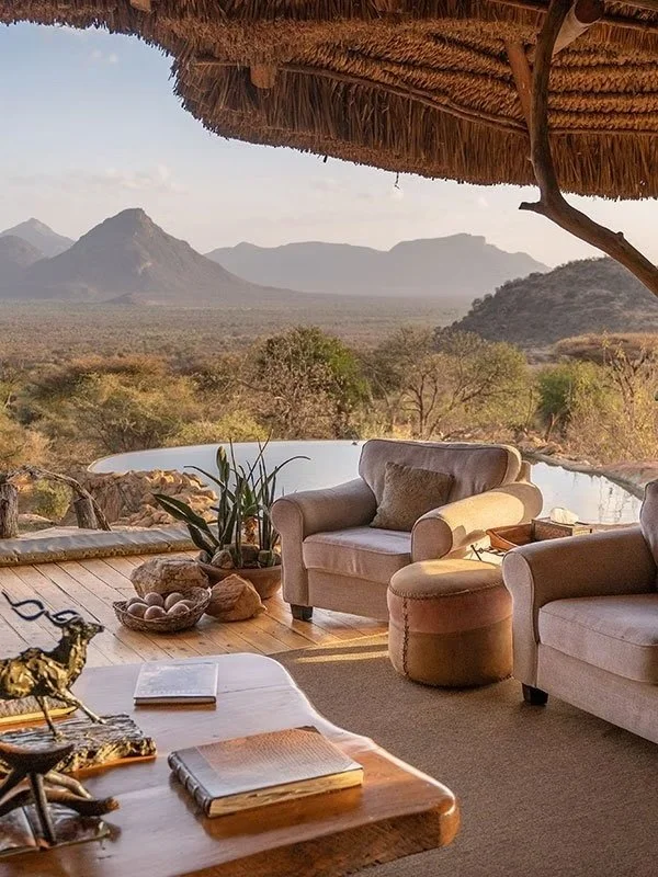 Living room with beige armchairs and a side table, overlooking a scenic landscape with mountains and trees, under a thatched roof.