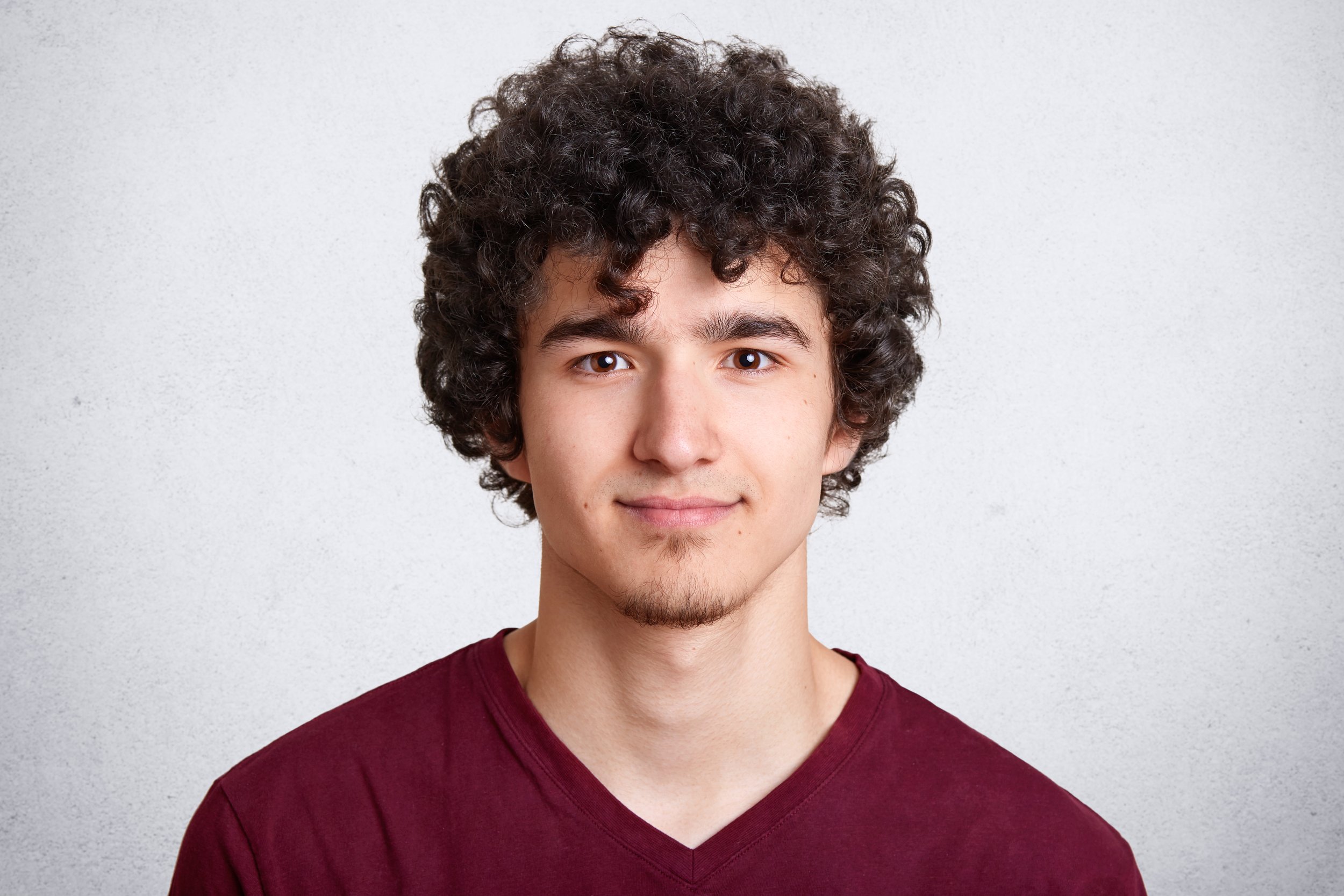 A young man with curly hair, light skin, and wearing a maroon shirt looking at the camera with a slight smile.