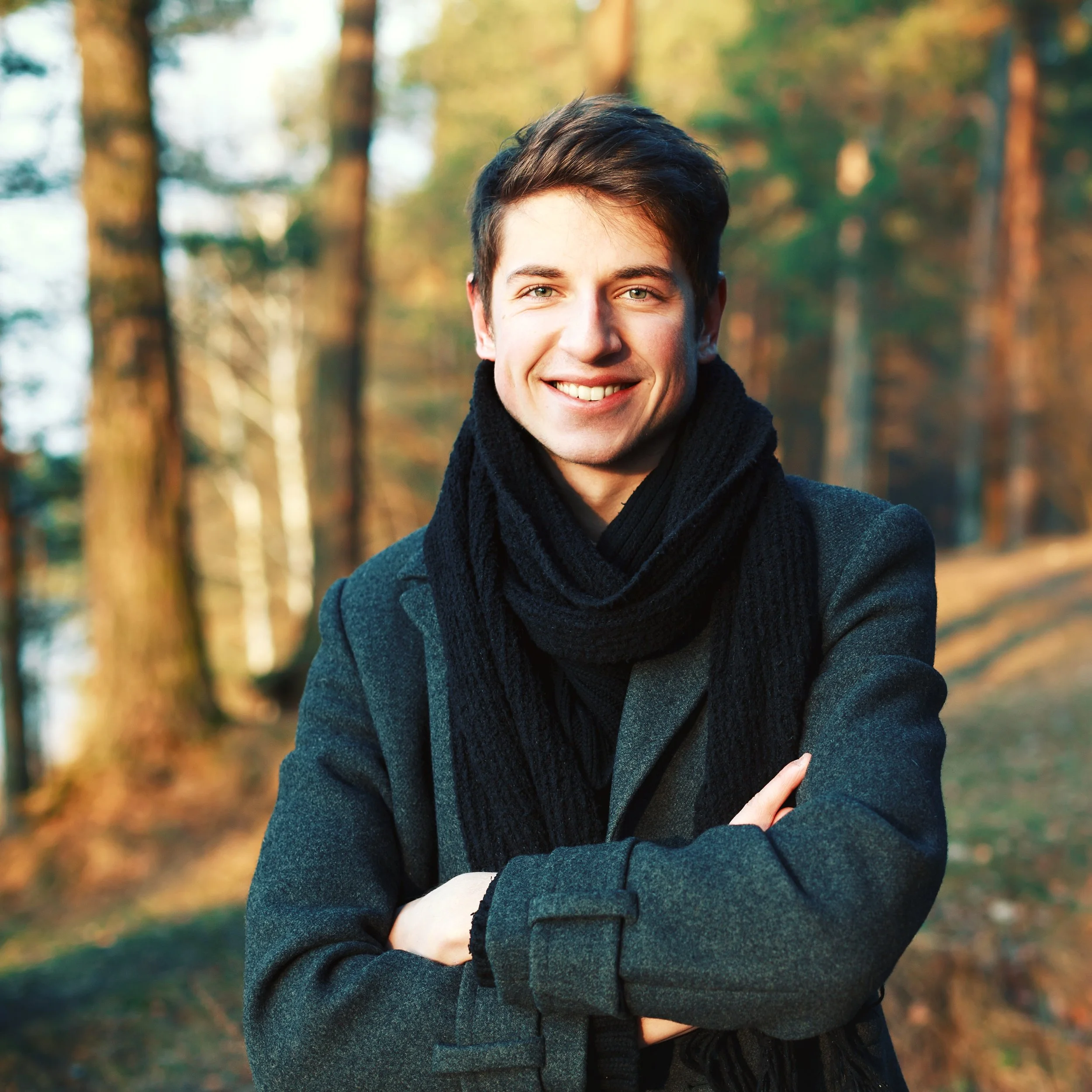 A young man smiling with arms crossed in a forest during autumn.