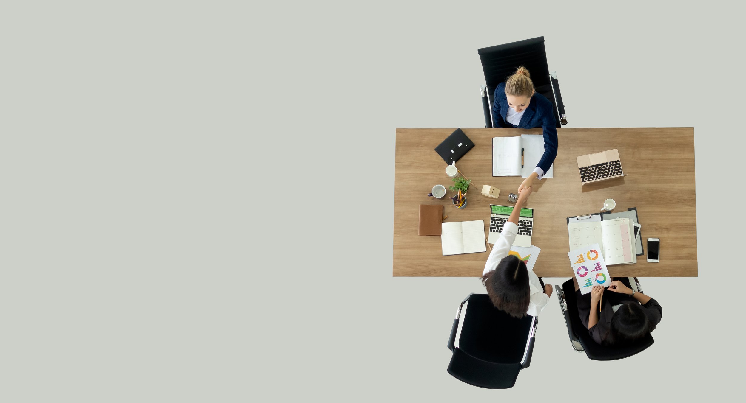 Overhead view of three people in formal business attire at a meeting, shaking hands over a wooden conference table with various documents, laptops, and office supplies.
