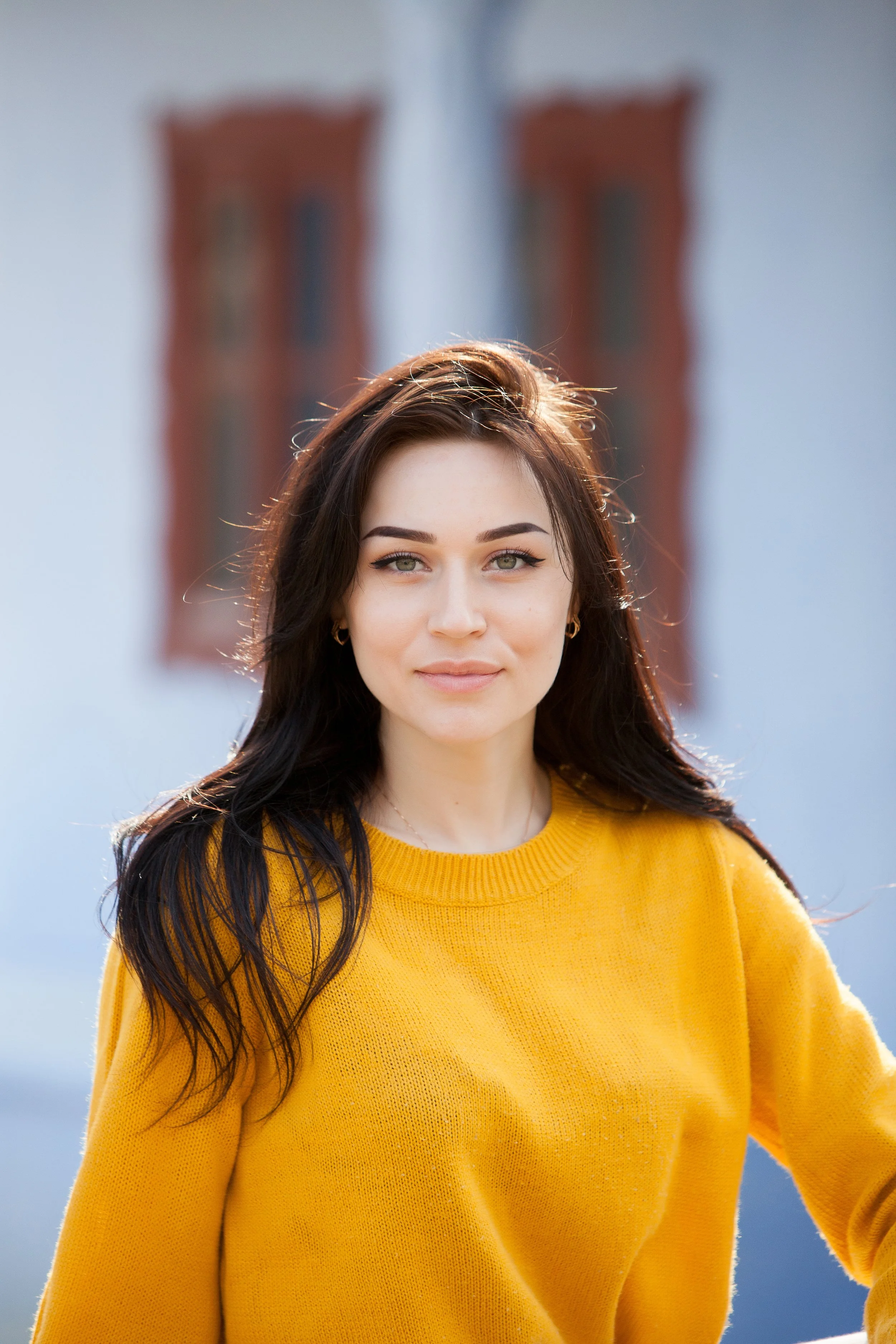 Young woman with dark hair and light skin wearing a yellow sweater outdoors, with a building and windows blurred in the background.