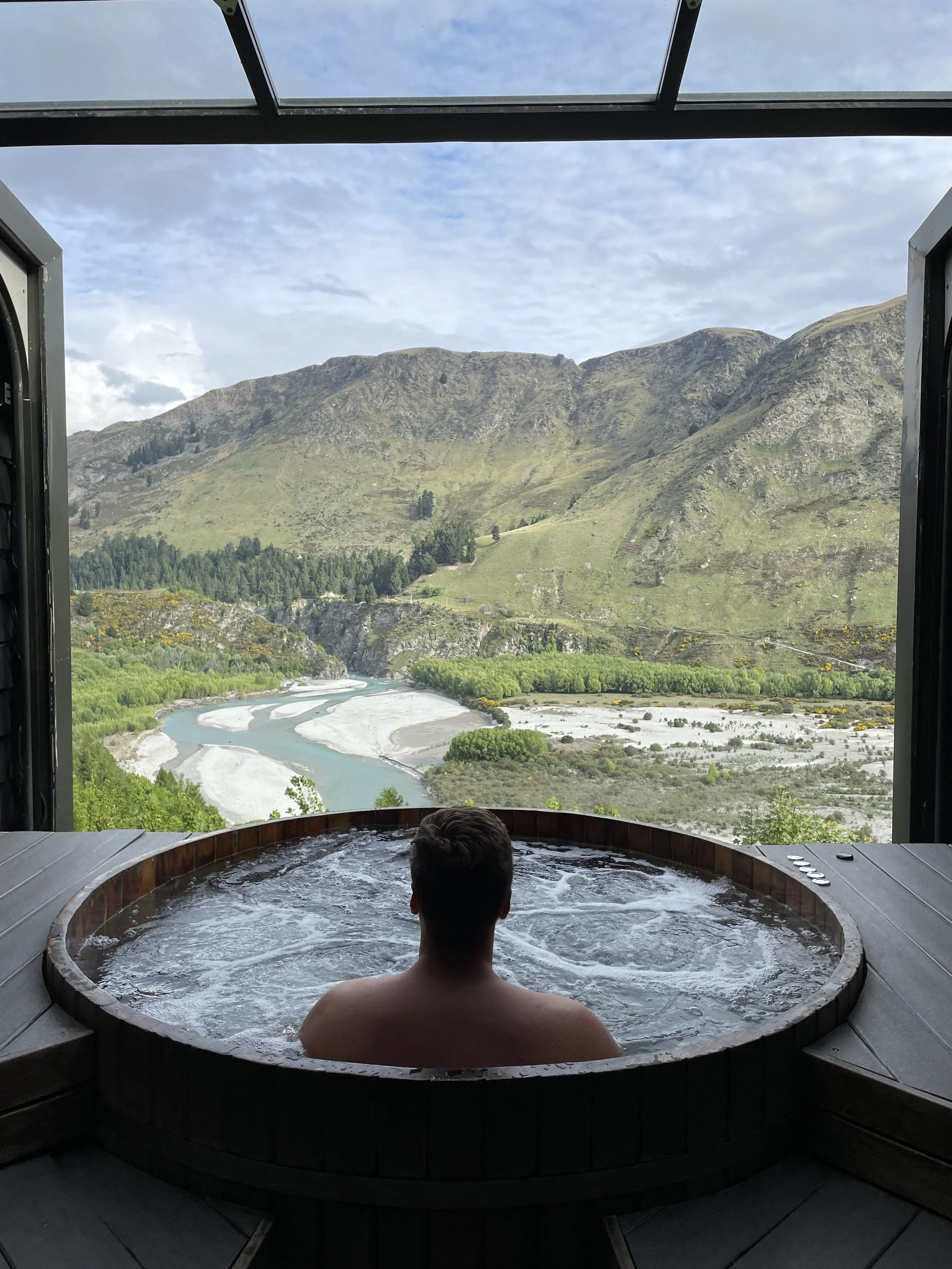 Person sitting in a hot tub overlooking a mountain landscape with a river and green trees