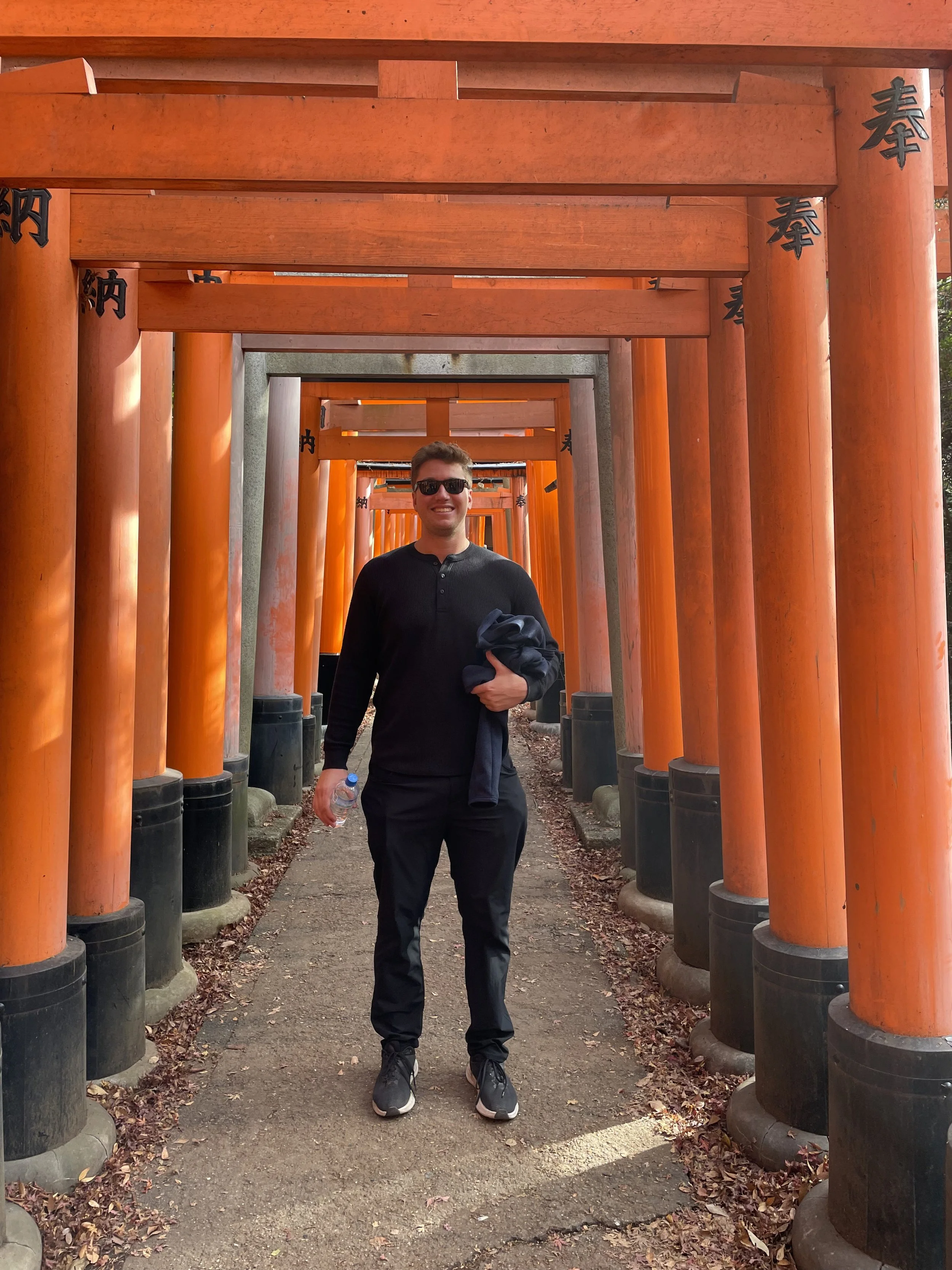 A man standing smiling in front of a series of red torii gates at Fushimi Inari Shrine in Japan, holding a water bottle.