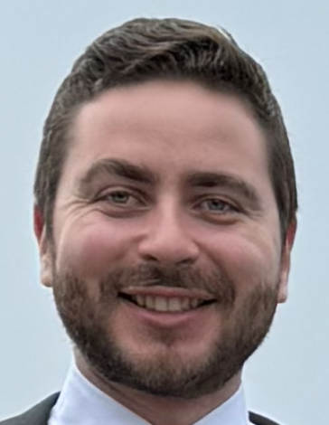 A young man with glasses and a beard standing outdoors near a car and a building, looking at the camera during sunset.
