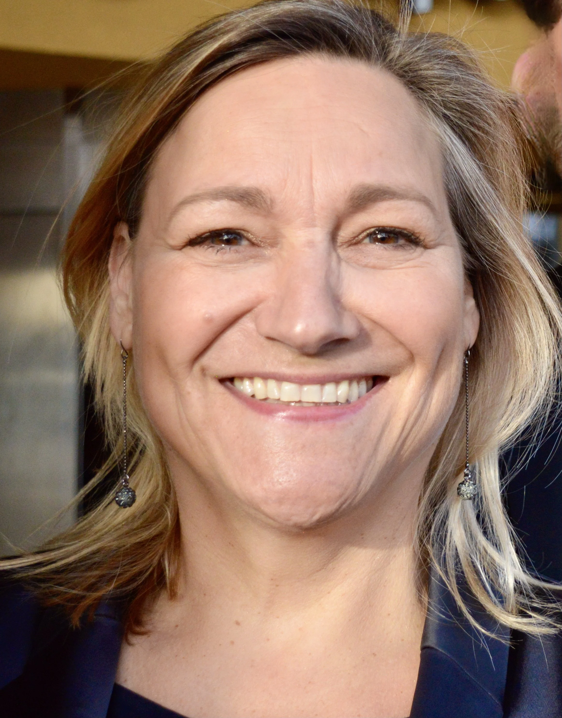 Close-up photo of a woman smiling with light makeup, wavy blonde hair, earrings, and a dark blazer.
