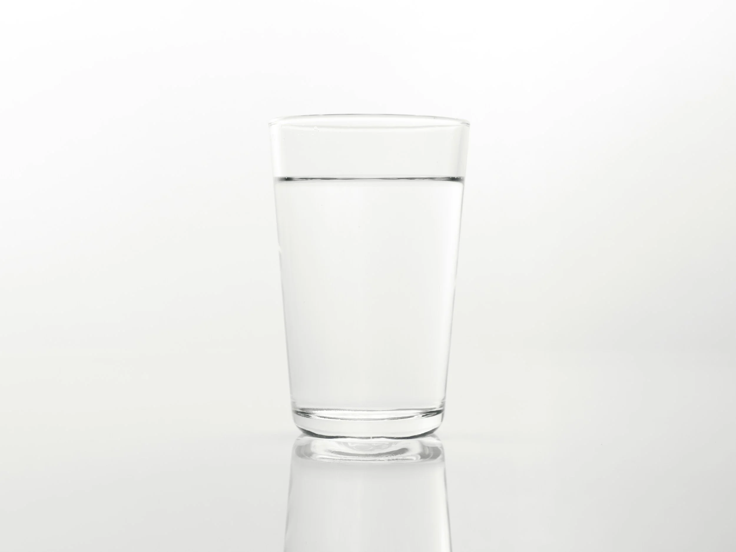 A clear glass of water on a white reflective surface.