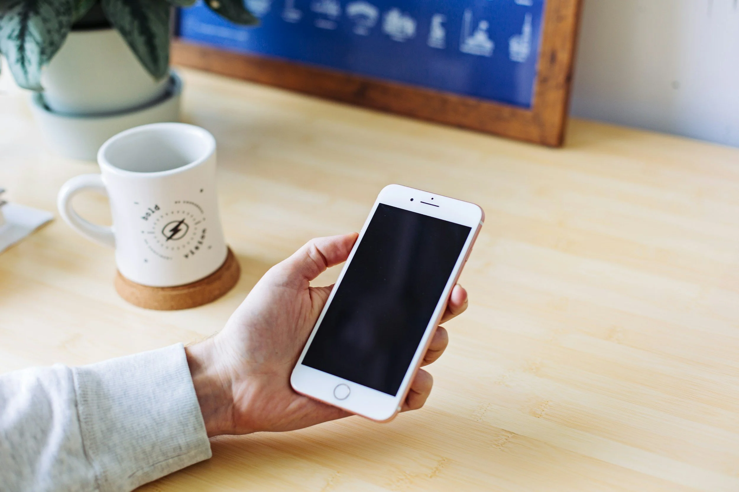 A person holding a white smartphone in their right hand, with a coffee mug and a potted plant on a wooden table. A framed blue print is in the background.