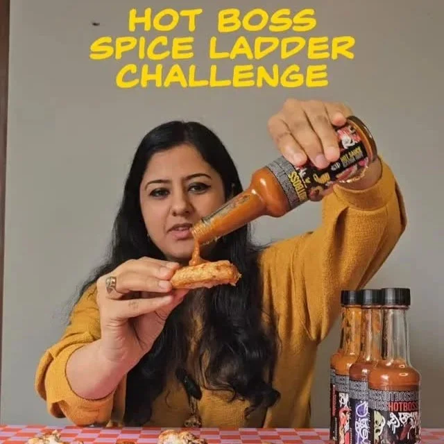 Woman wearing a mustard yellow sweatshirt holds a slice of pizza in one hand and pours hot sauce from a bottle onto it with her other hand, with a jar of hot sauce on the table, and a plain gray background. Bright yellow text reads 'HOT BOSS SPICE LADDER CHALLENGE' at the top.