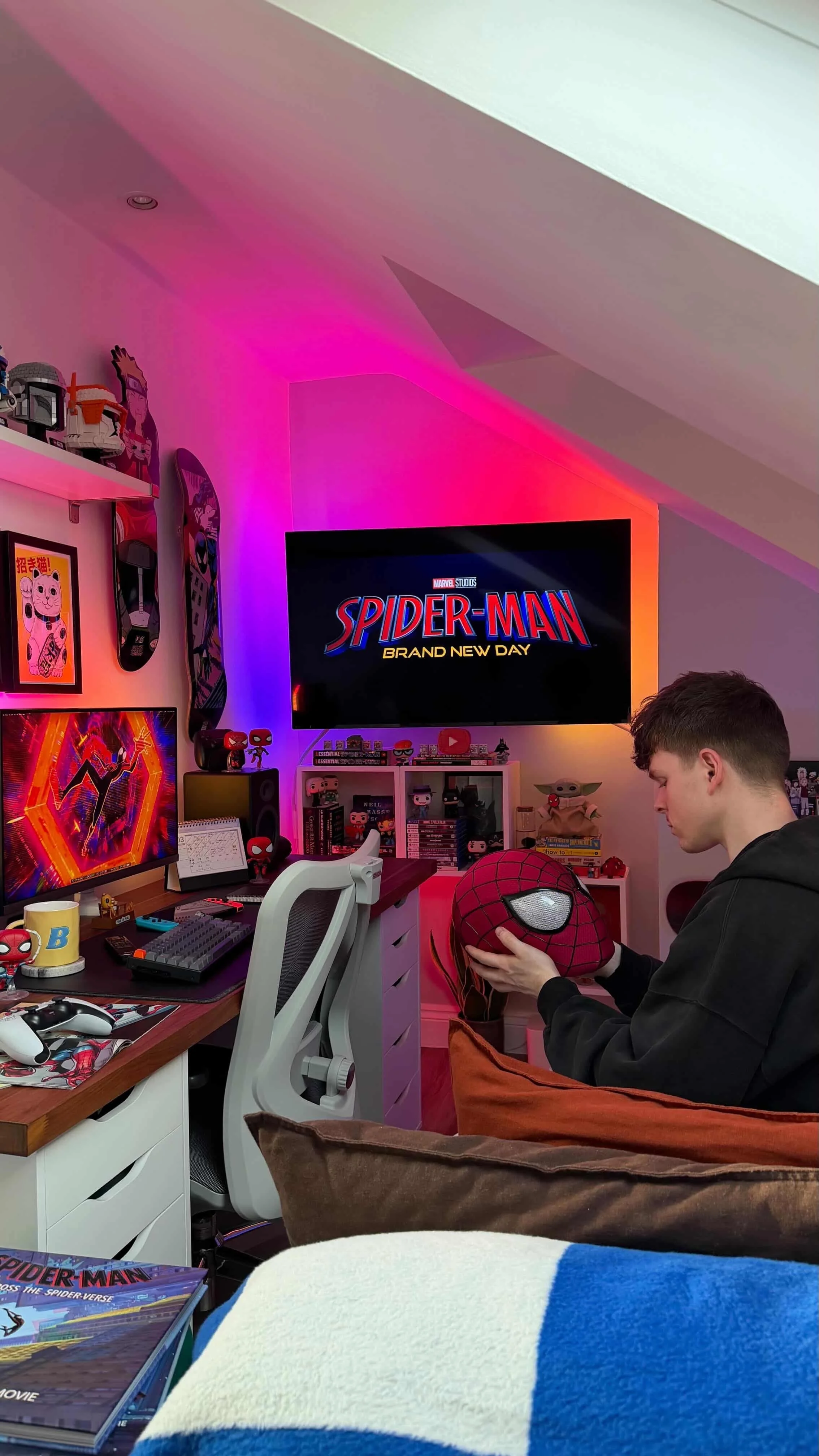 A person sitting in a room decorated with Spider-Man items, holding a Spider-Man pillow, with a large TV displaying the "Spider-Man: Brand New Day" logo. The room has colorful lighting, and there are various figurines, wall art, and gaming accessories on the desk and shelves.