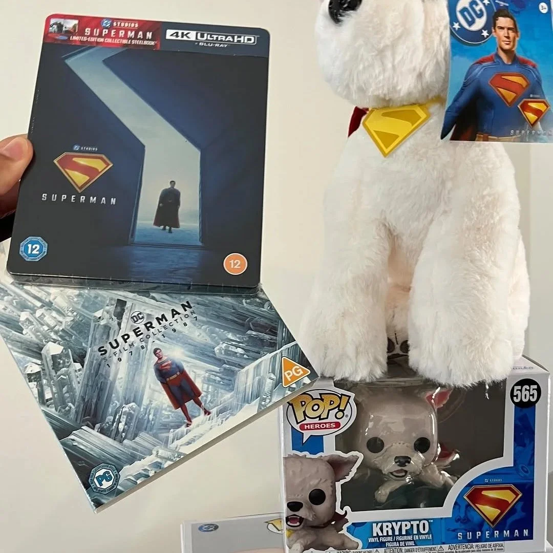 Superman themed collectibles including a Blu-ray box set, a plush dog wearing a Superman collar, a Funko Pop figure, and a comic book, all related to Superman.