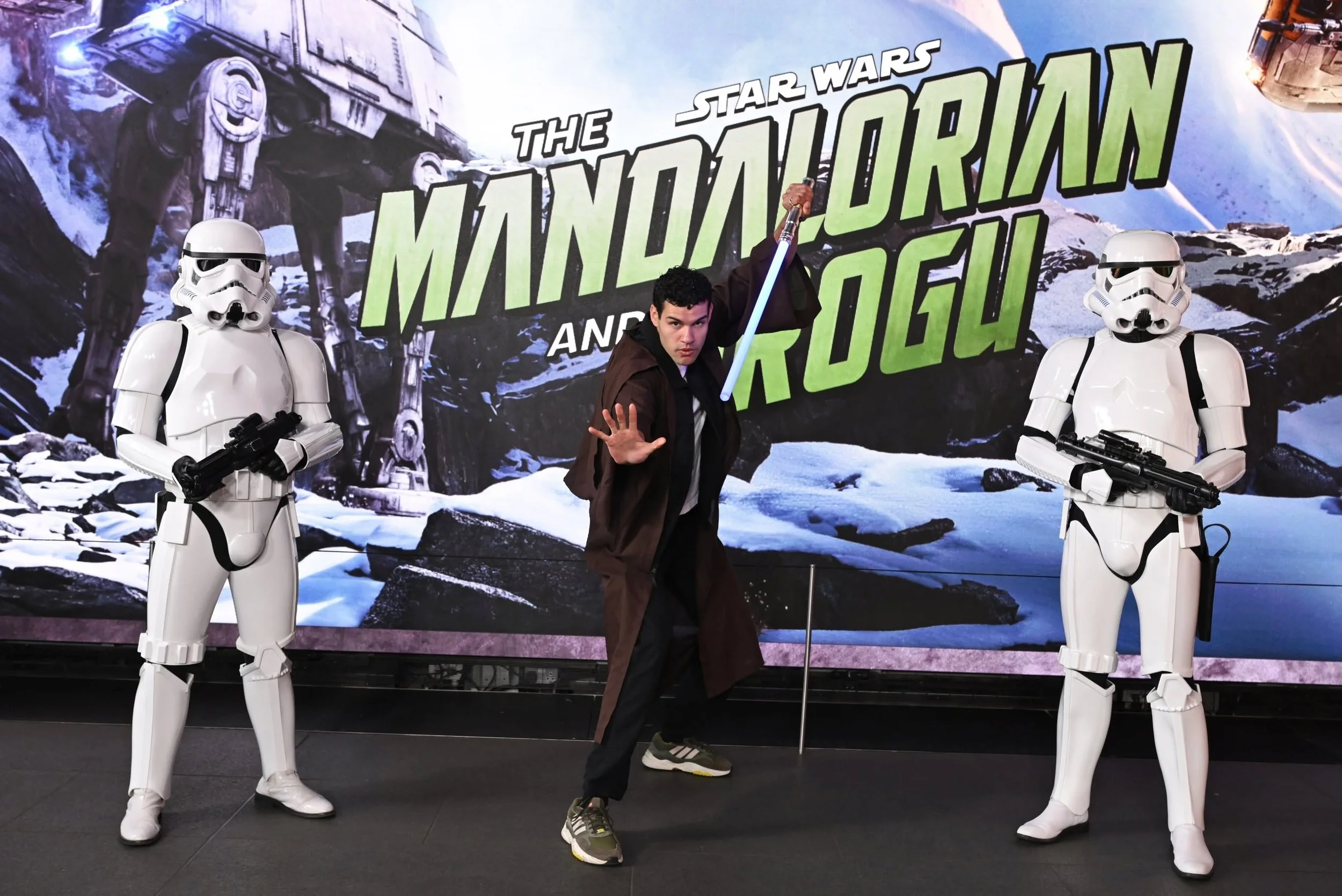 A person holding a lightsaber pose between two Stormtroopers in front of a Star Wars themed backdrop.
