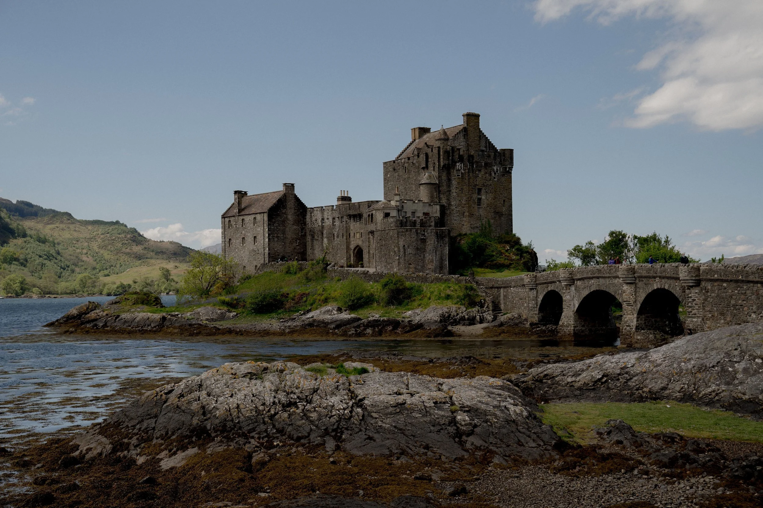 A stone castle with a bridge over a river, surrounded by green hills and a cloudy sky.