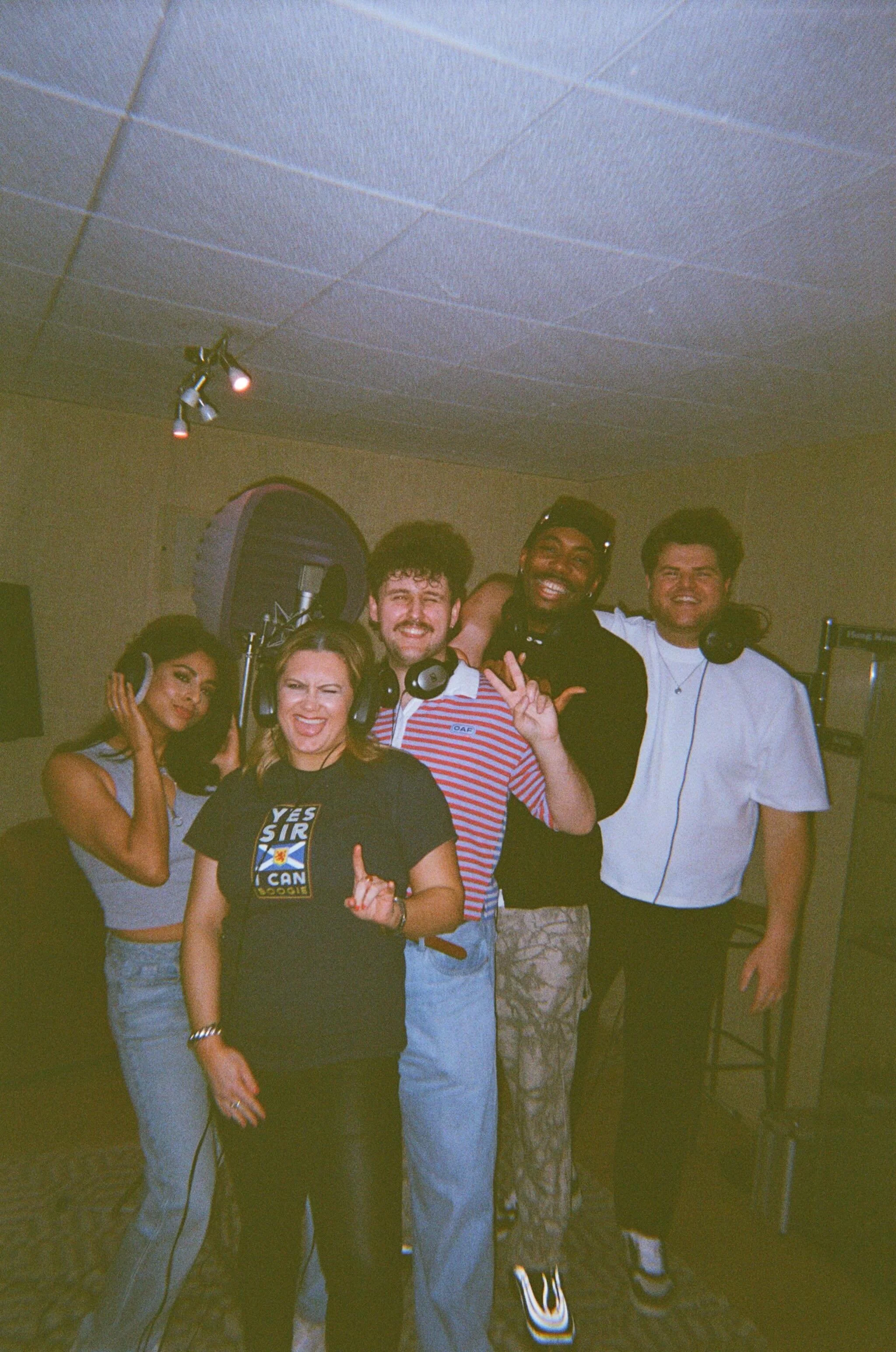 A group of six young adults inside a recording studio, some wearing headphones, smiling and posing for the photo.