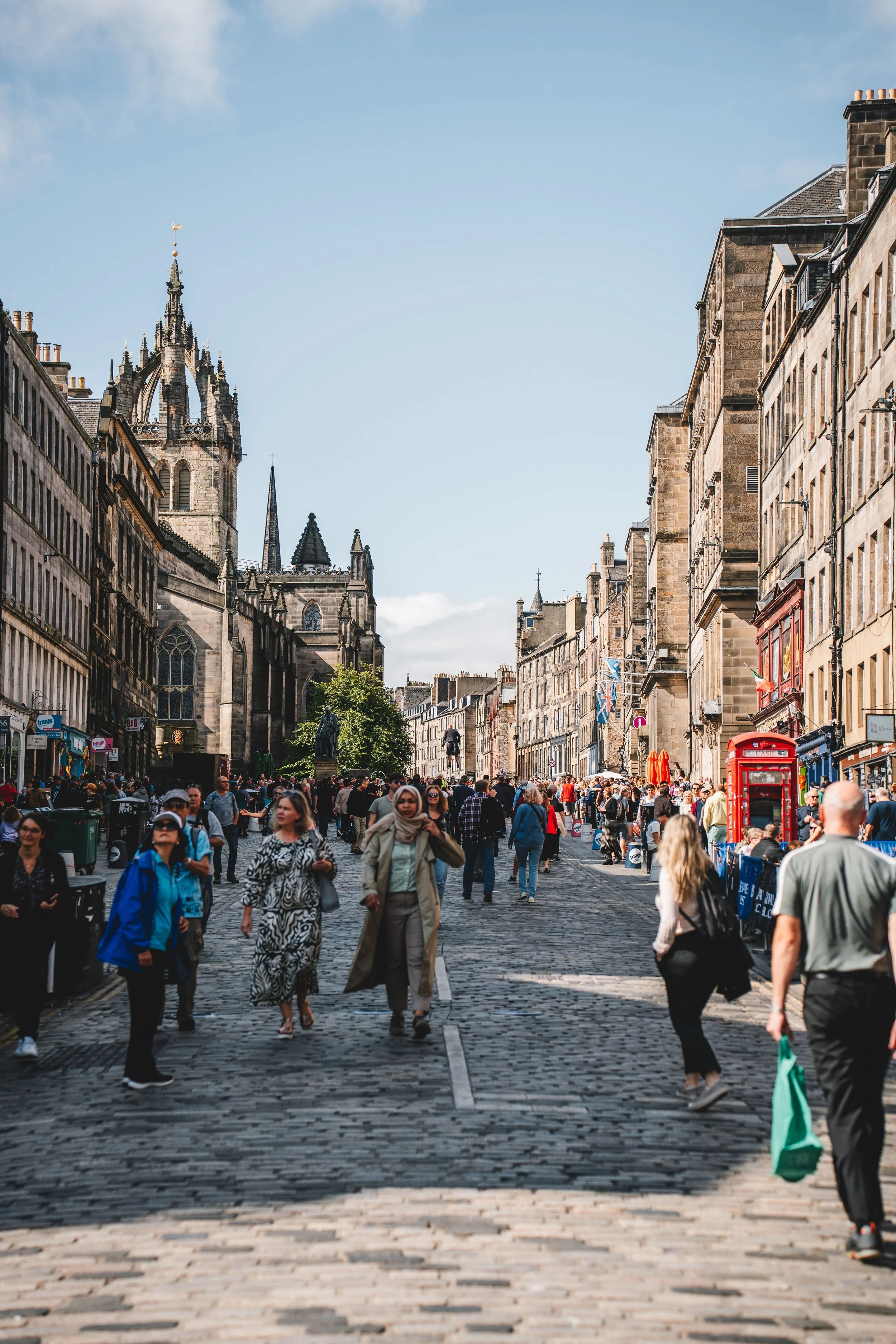 A busy city street with pedestrians walking on cobblestone pavement, historic buildings with flags, and a large church with tall spires in the background under a partly cloudy sky.
