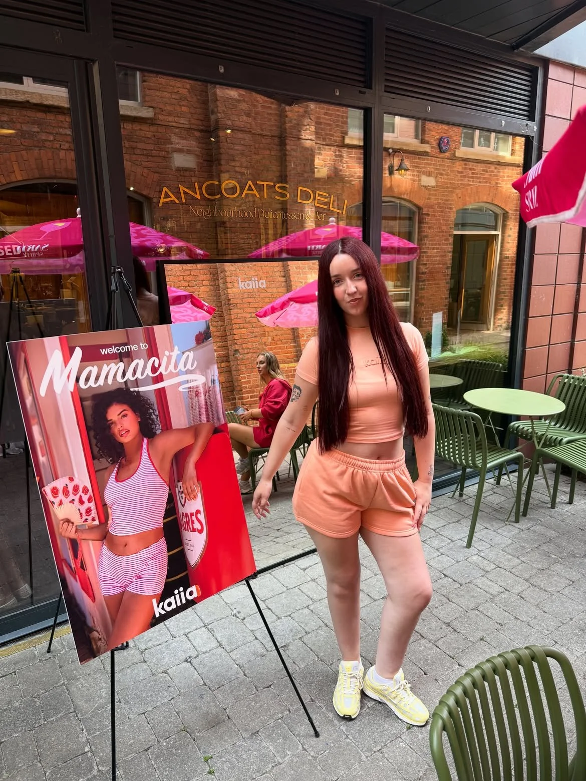 A young woman with long dark hair wearing a peach-colored outfit standing outside a cafe with green chairs and tables, a large promotional poster of another woman dressed in red and white stripes, and pink umbrellas in the background.