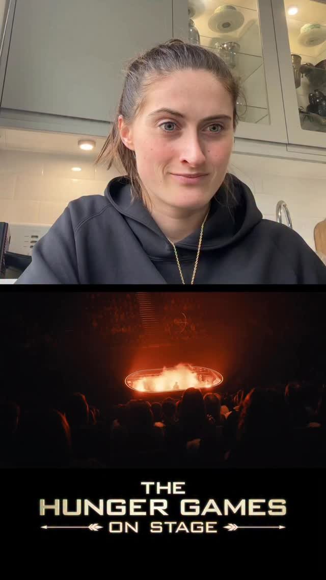 A woman with brown hair tied back in a ponytail, wearing a black hoodie and a gold necklace, sitting at a kitchen counter. In the lower part of the image, there is a scene from 'The Hunger Games' movie showing a fiery arena with an audience watching, along with the movie's logo that reads 'The Hunger Games on Stage'.