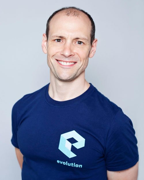 A smiling man with short brown hair, wearing a navy blue t-shirt with a logo and the word "evolution" on it, standing against a light gray background.