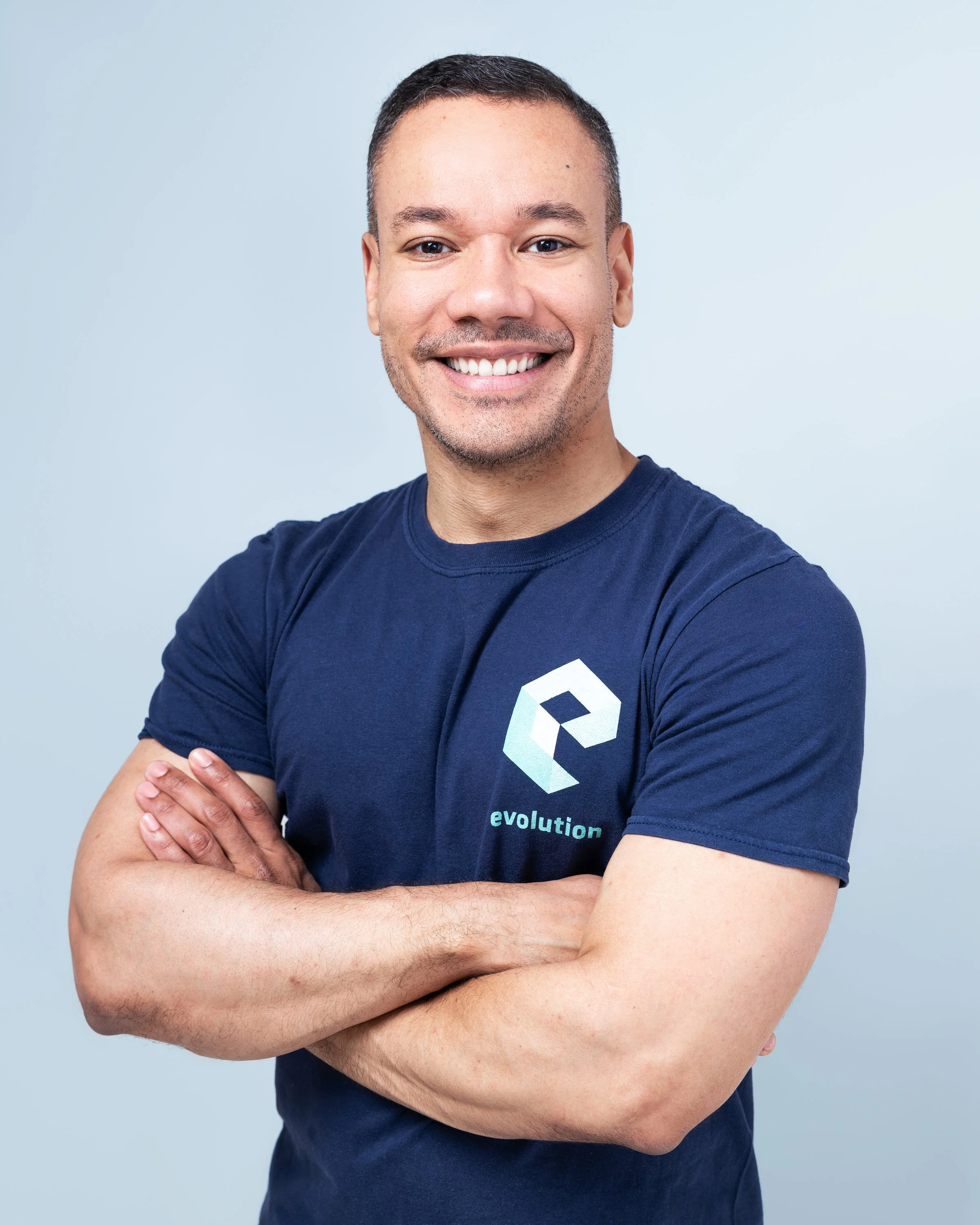 A smiling man with short dark hair wearing a navy blue t-shirt with a geometric logo and the word 'evolution' on it, standing with arms crossed against a light blue background.