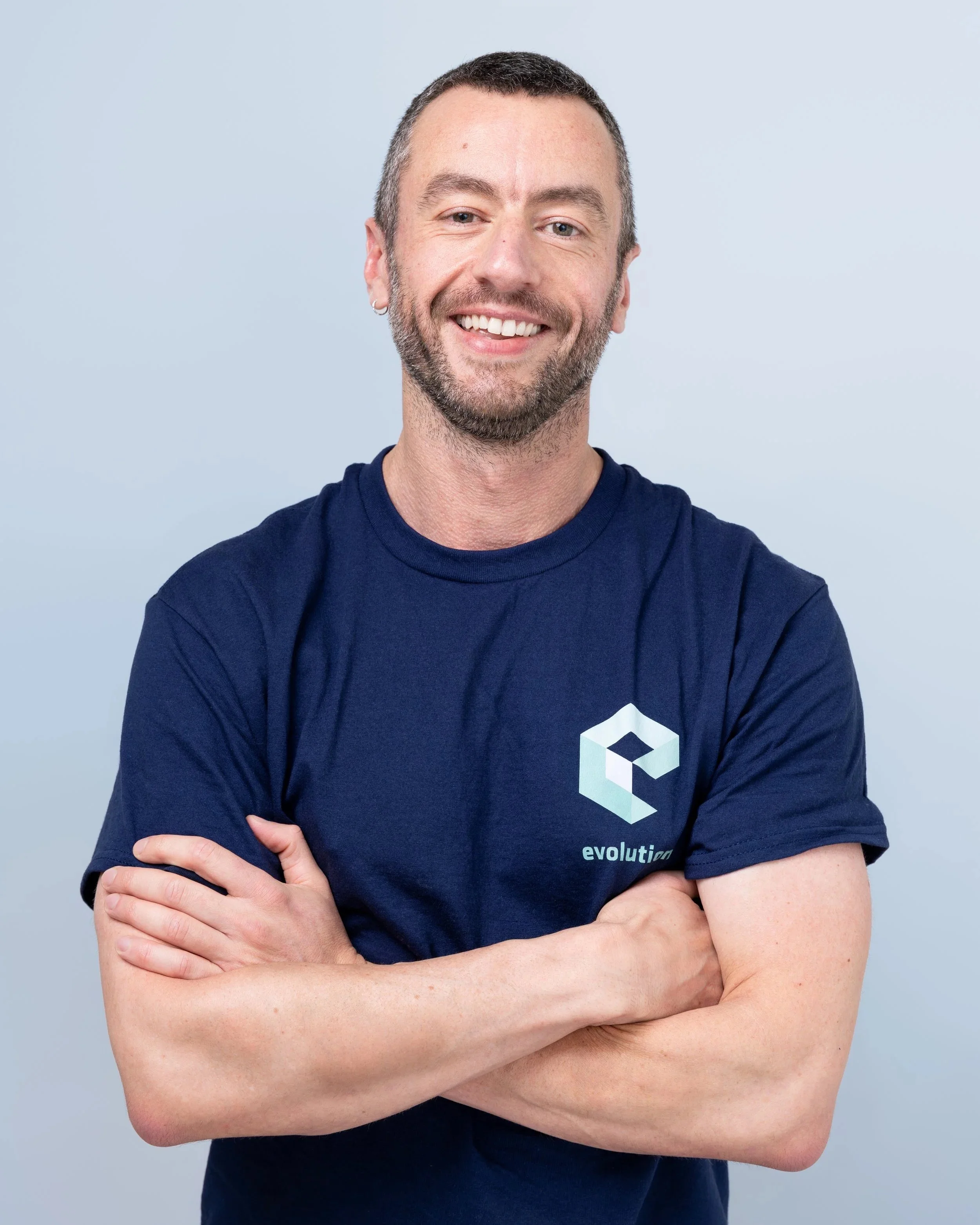 A smiling man with short dark hair, a beard, and a tattoo on his left arm, wearing a navy blue T-shirt with a geometric logo and the word 'evolution' on it, standing with arms crossed against a plain light gray background.