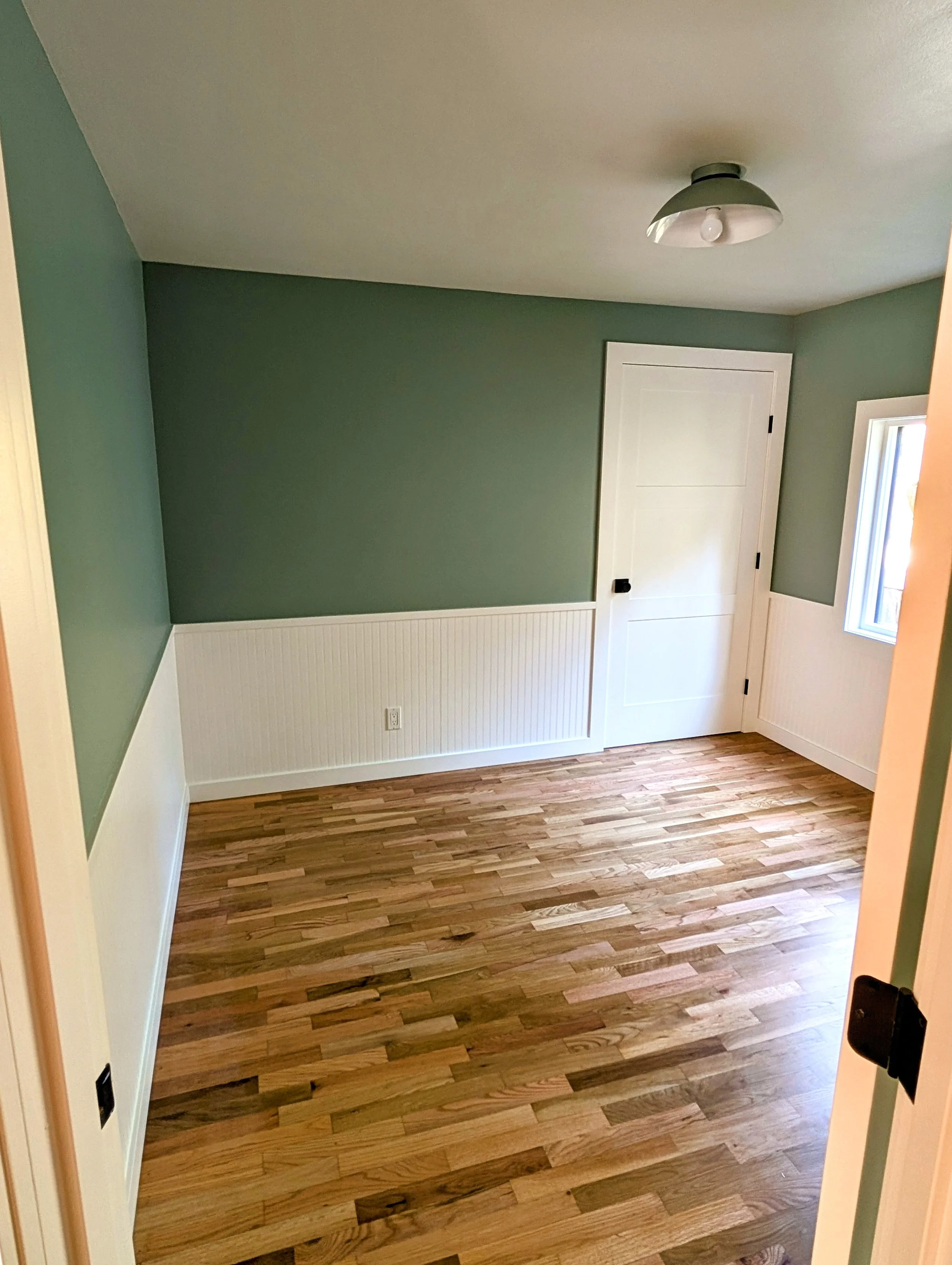 Empty room with green upper walls, white wainscoting, hardwood floor, window, and ceiling light fixture.