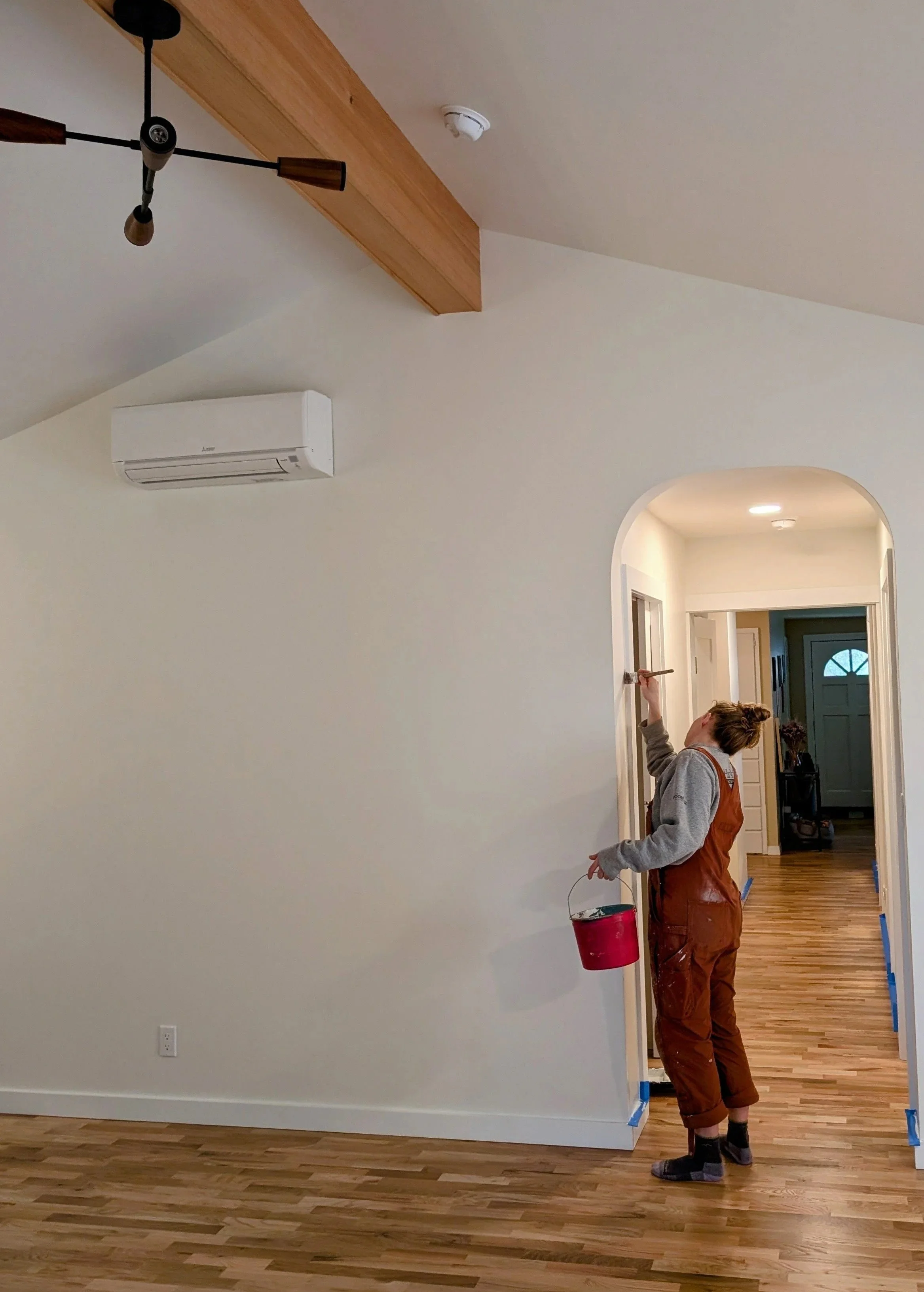 A woman painting a wall in a hallway with a paintbrush and holding a red paint bucket, wearing brown overalls and gray sweatshirt, with hardwood floors and a white wall, with an air conditioning unit on the wall and a modern black ceiling light fixture.