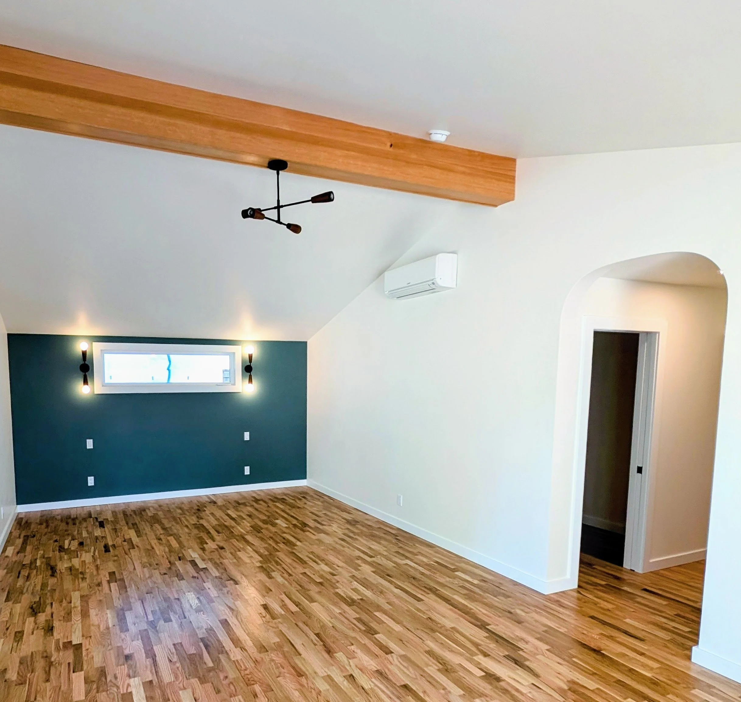 Empty living room with hardwood floors, a teal accent wall with a horizontal window, sliding glass door, modern black chandelier, and a wall-mounted air conditioning unit.