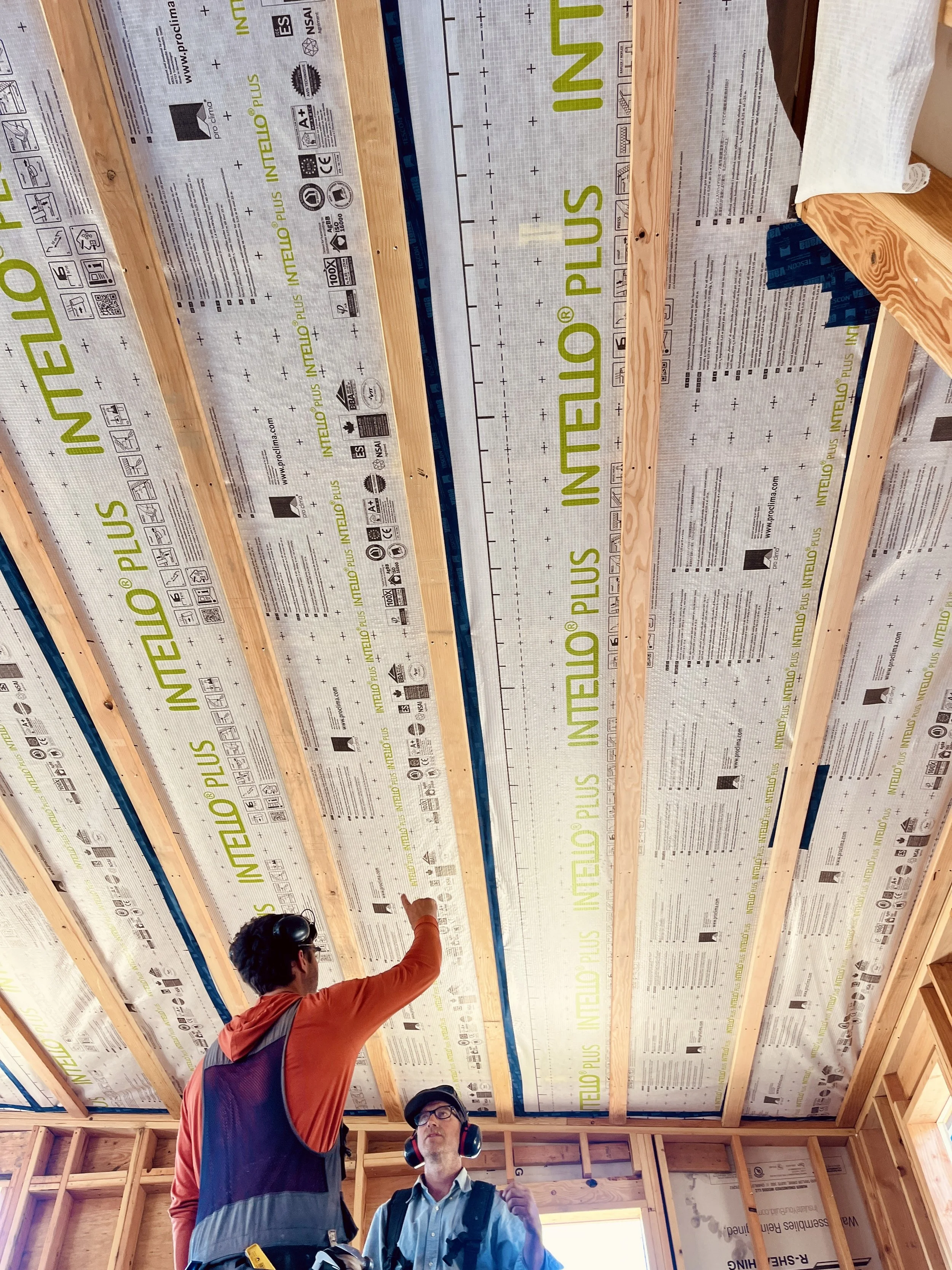 Two construction workers inside a building under construction, with visible wooden beams and insulation, one pointing at the ceiling and the other listening, both wearing safety gear.