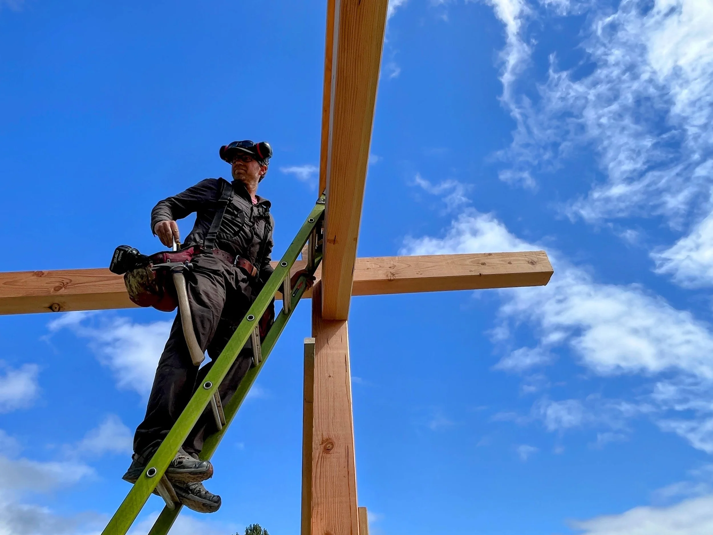 A construction worker on a ladder working on a wooden frame against a blue sky with clouds.