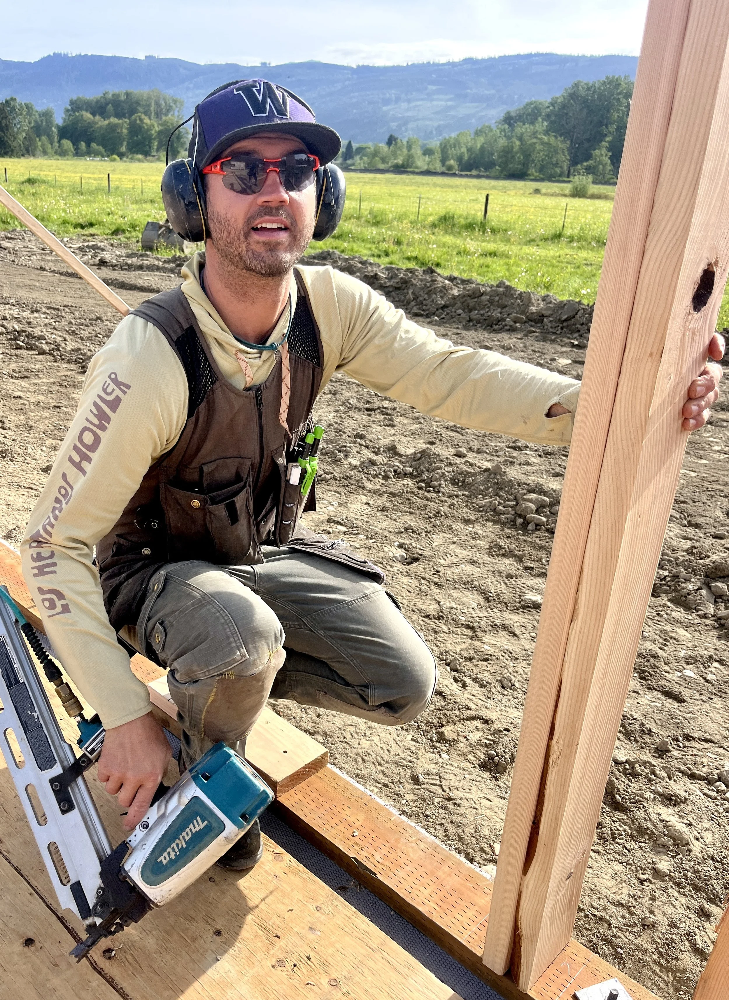 A construction worker wearing a cap, glasses, ear protection, and a beige long sleeve shirt kneeling on a wooden platform, holding a nail gun, and working on a wooden frame outdoors with a grassy field and hills in the background.