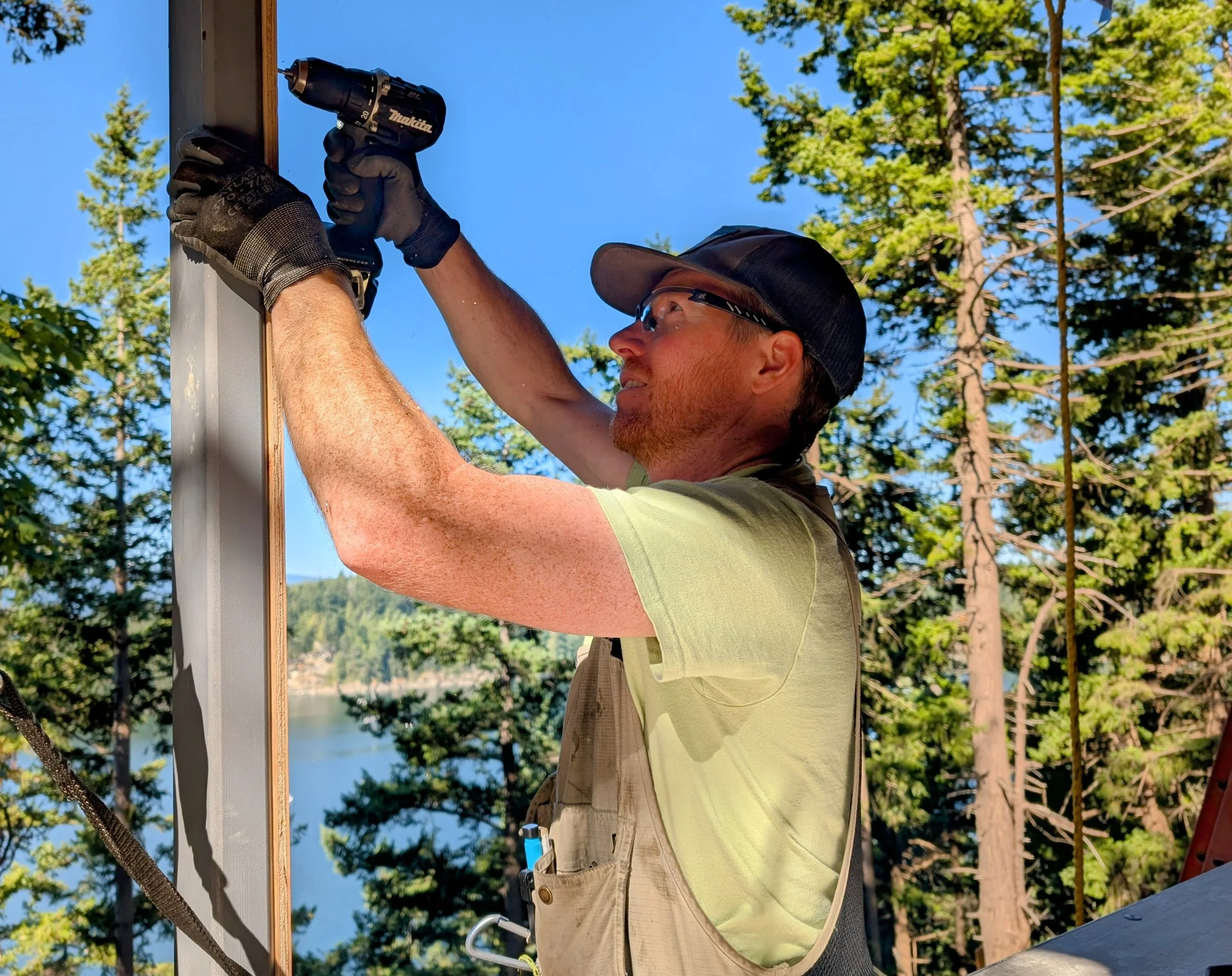 A man wearing safety glasses, a baseball cap, and work gloves is using a power drill to attach a wooden board to a vertical metal post outdoors, with trees and a body of water visible in the background.