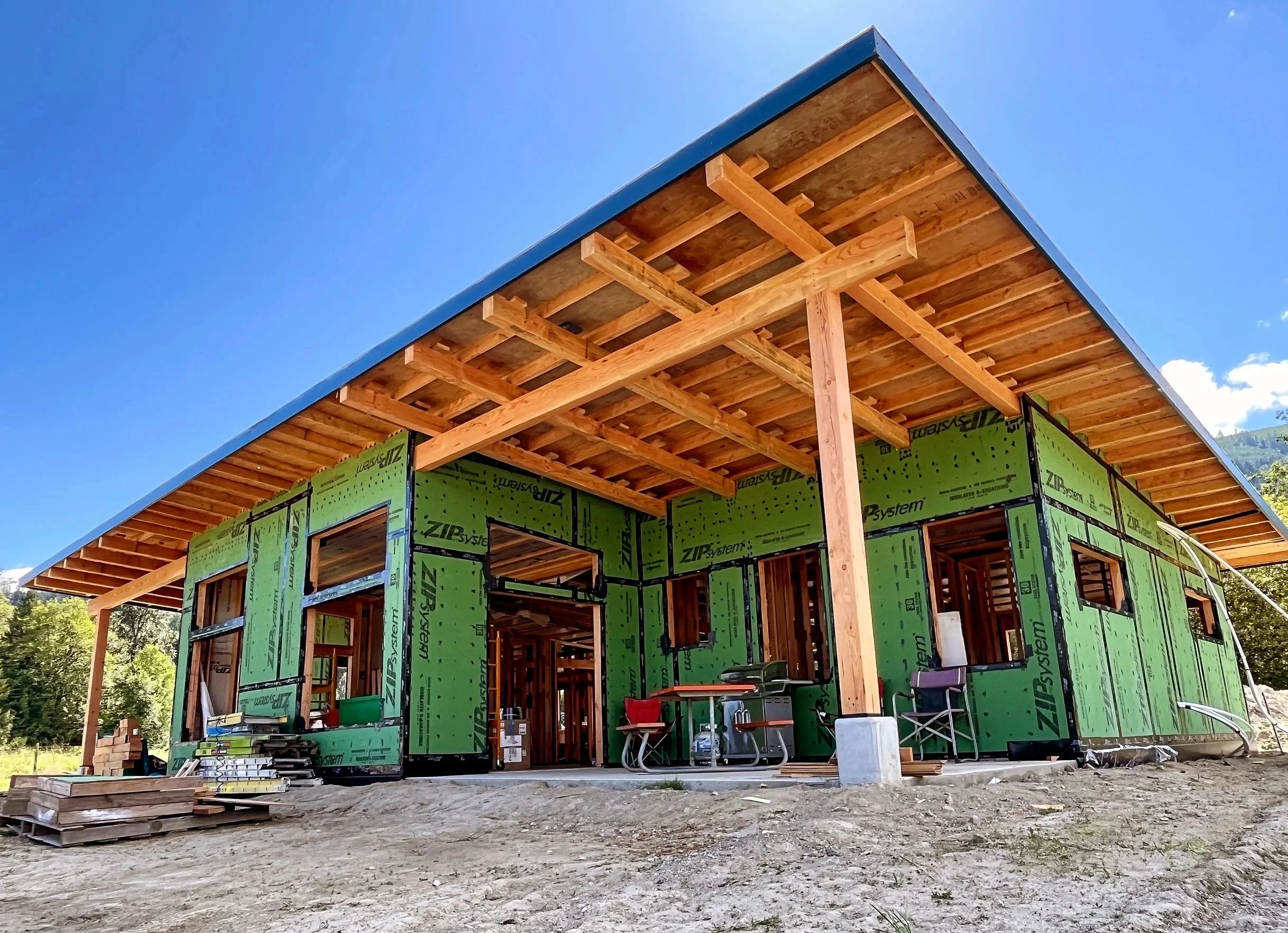 Under construction house with green weather-resistant sheathing, exposed wooden framing, and a large overhanging roof, set against a bright blue sky and distant trees.