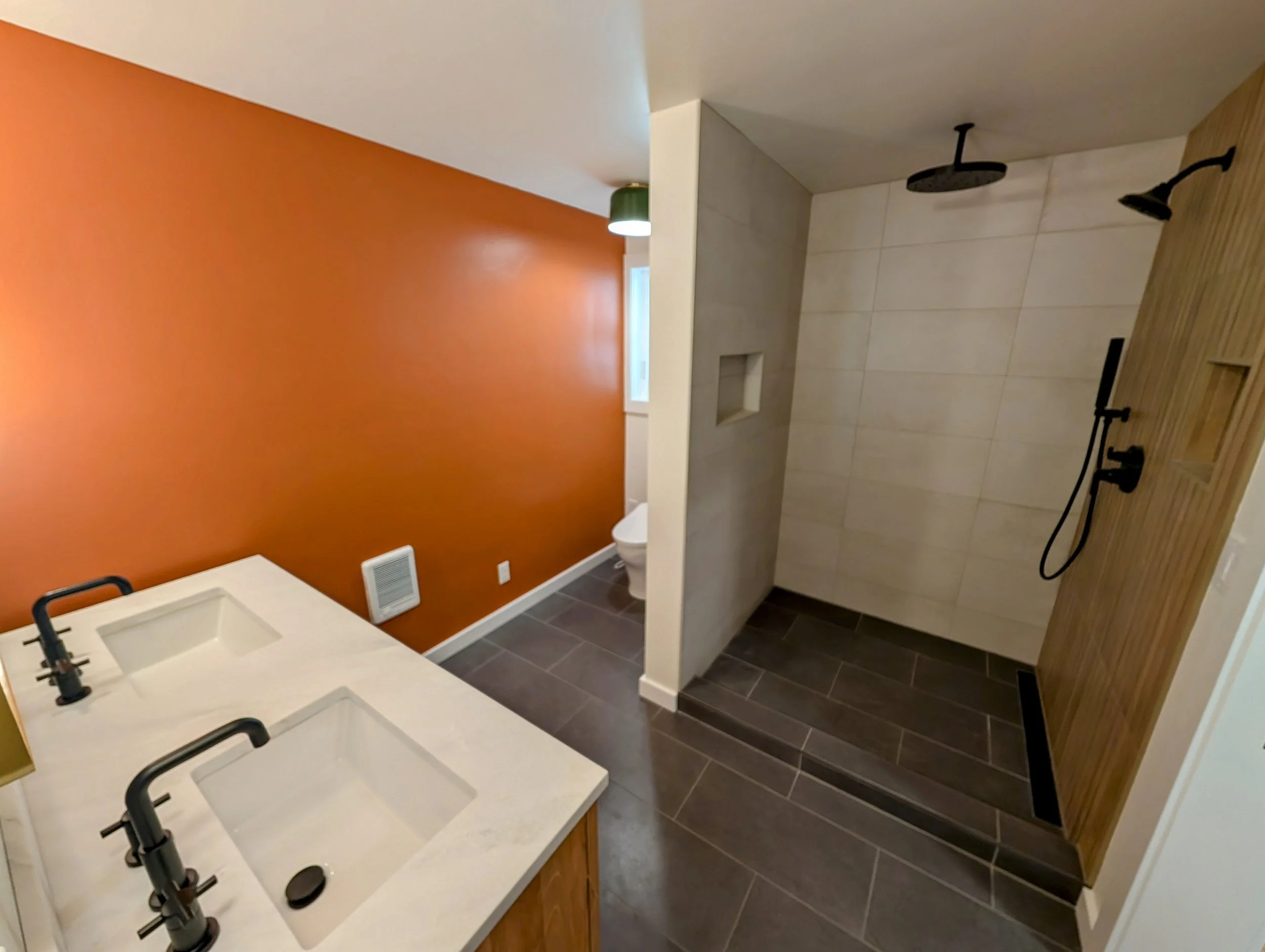 Bathroom with double vanity sink, standing shower with black fixtures, orange accent wall, gray floor tiles, and white toilet.