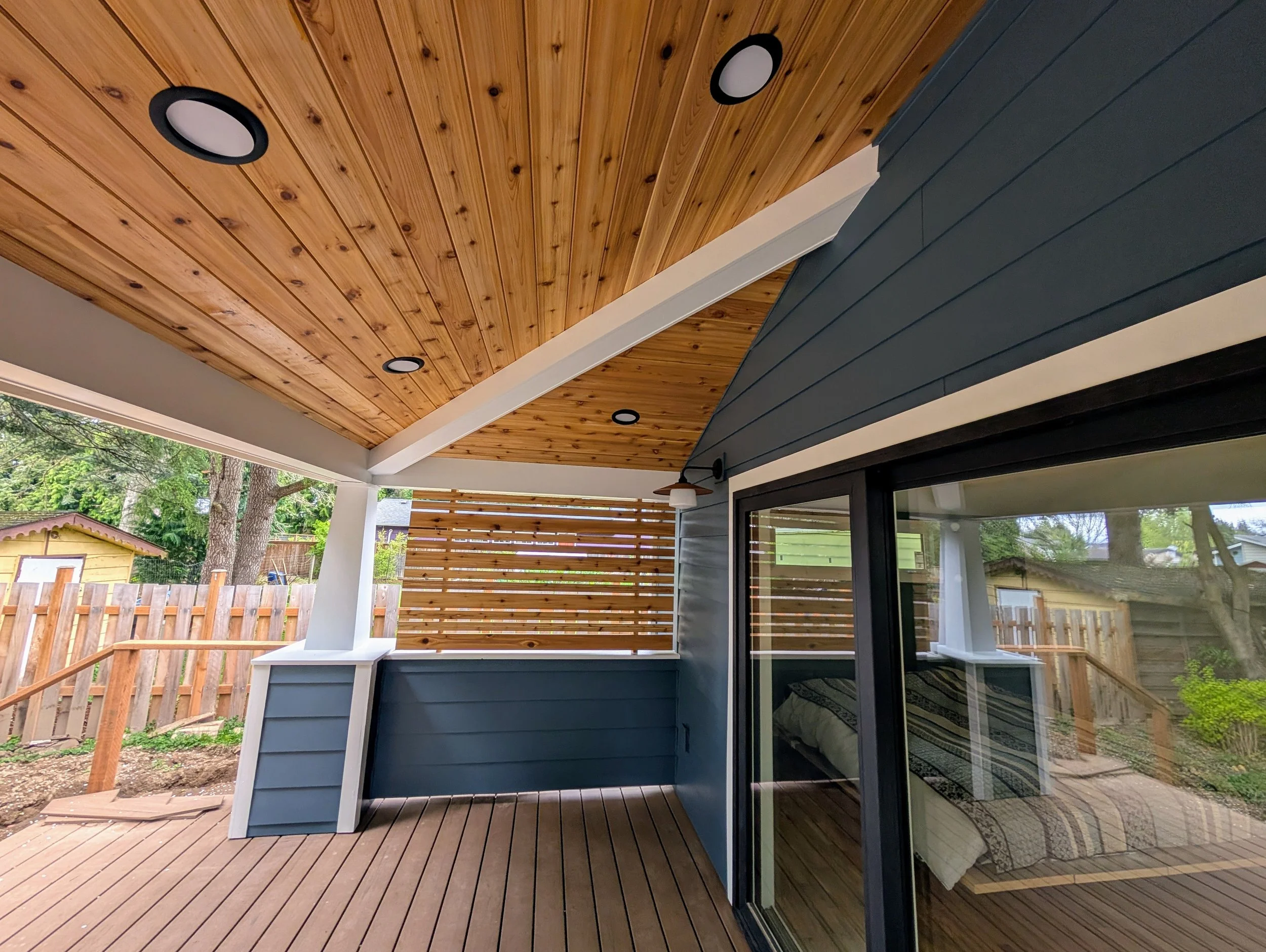 Outdoor covered deck with wood flooring and ceiling, black recessed lighting, blue exterior wall with large glass sliding door, wooden privacy screen, and backyard with trees and a small yellow shed.