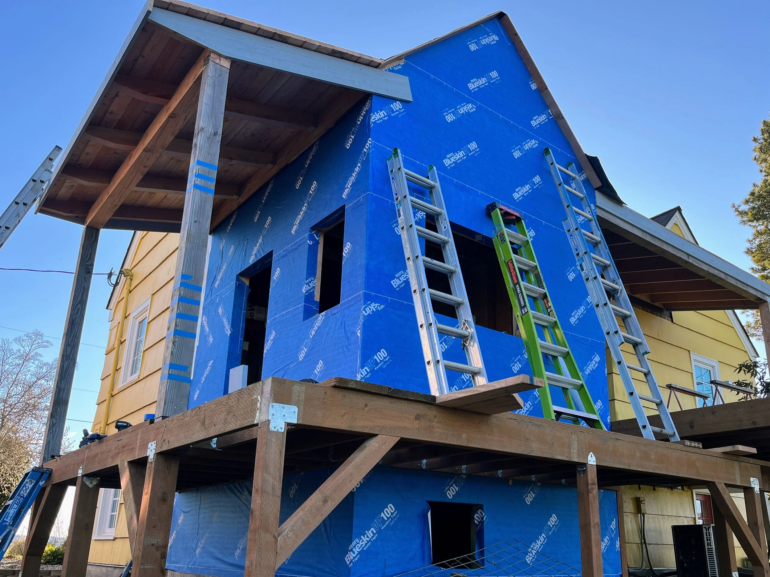 Front porch under construction with blue insulation board on walls, a wooden door, a window, a ladder, and construction tools.