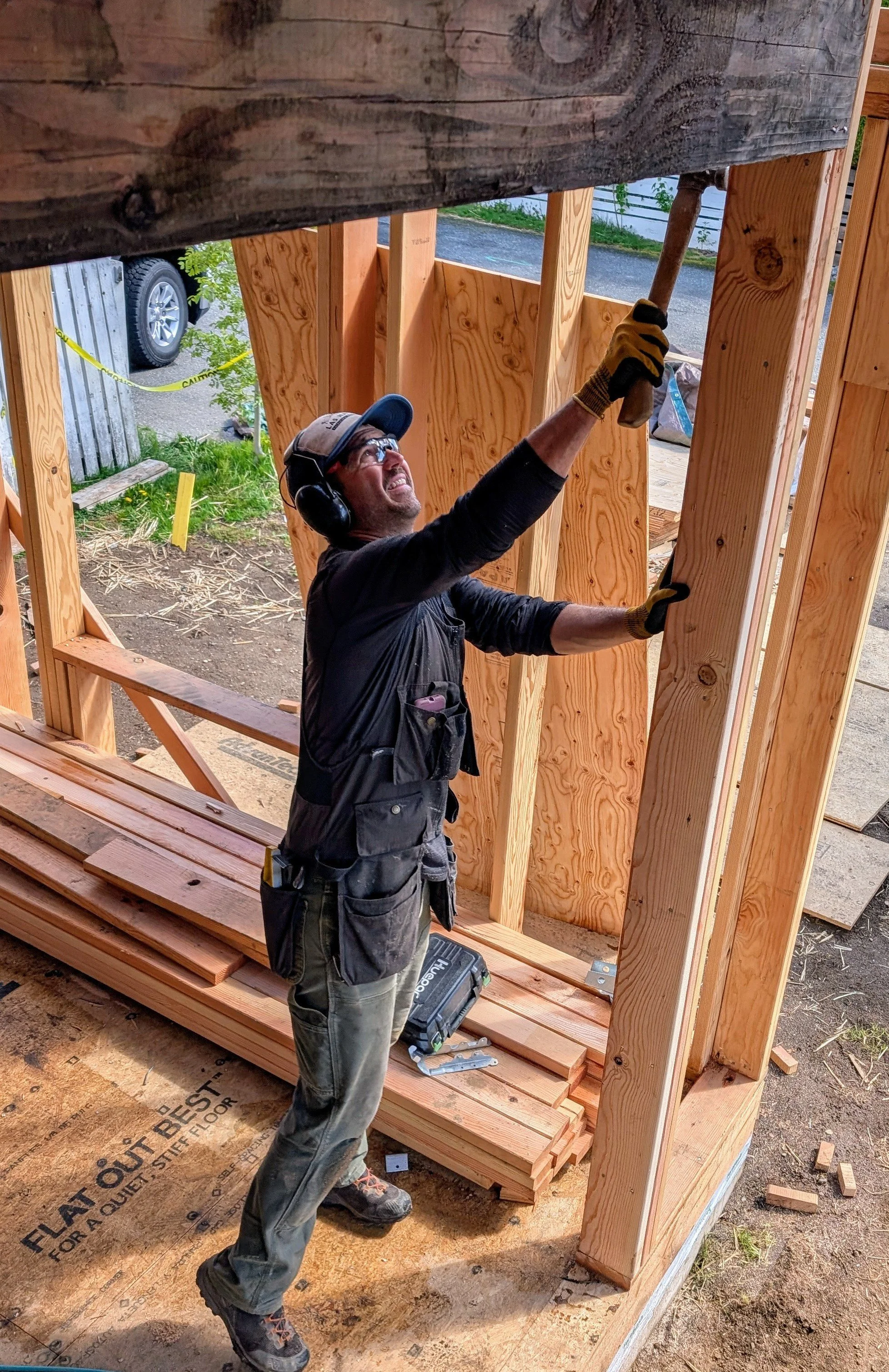 A construction worker wearing safety goggles, gloves, and a tool belt is using a hammer to nail a wooden beam during building.