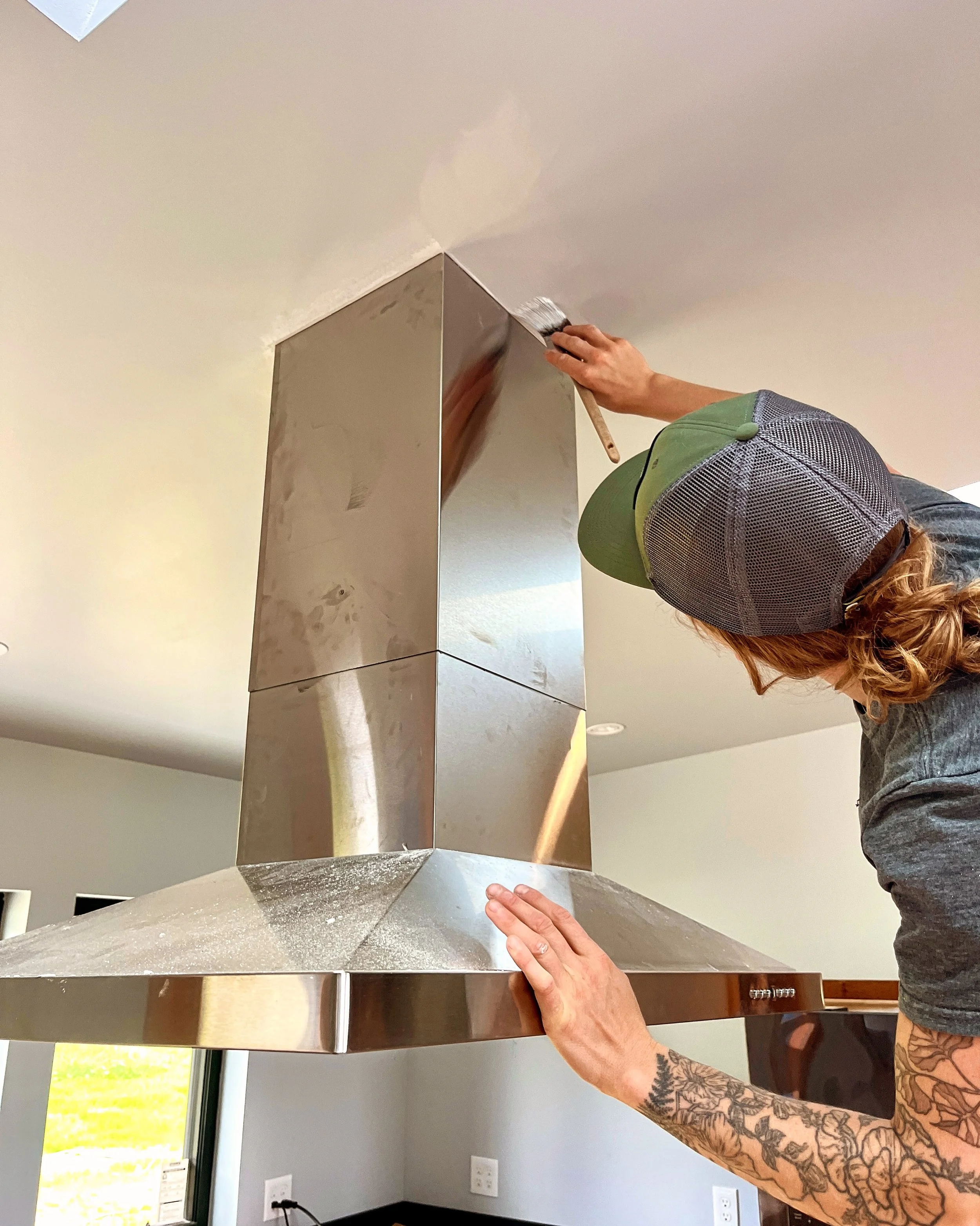 Person with a tattooed arm and a green and gray cap cleaning a stainless steel kitchen range hood with a brush.