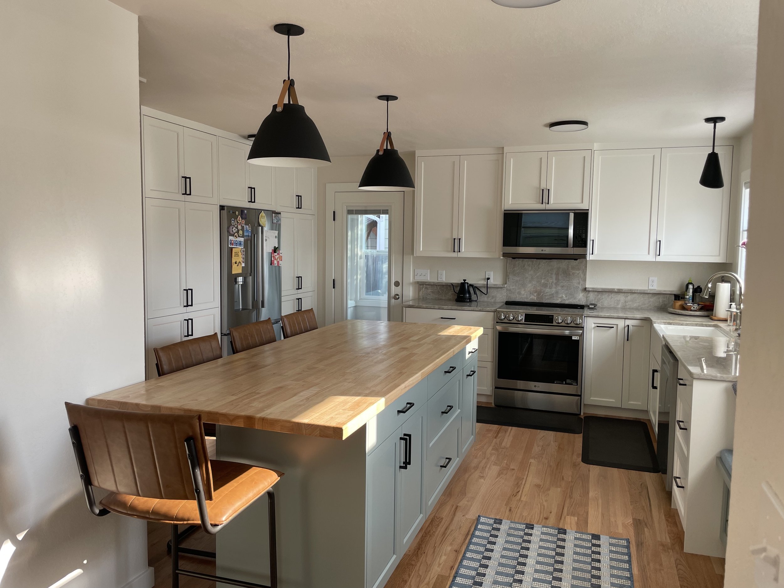 Modern kitchen with white cabinets, black hardware, a stainless steel refrigerator, oven, microwave, and a kitchen island with a wooden countertop. There are three black pendant lights above the island, a small rug on the wooden floor, and a door leading outside.