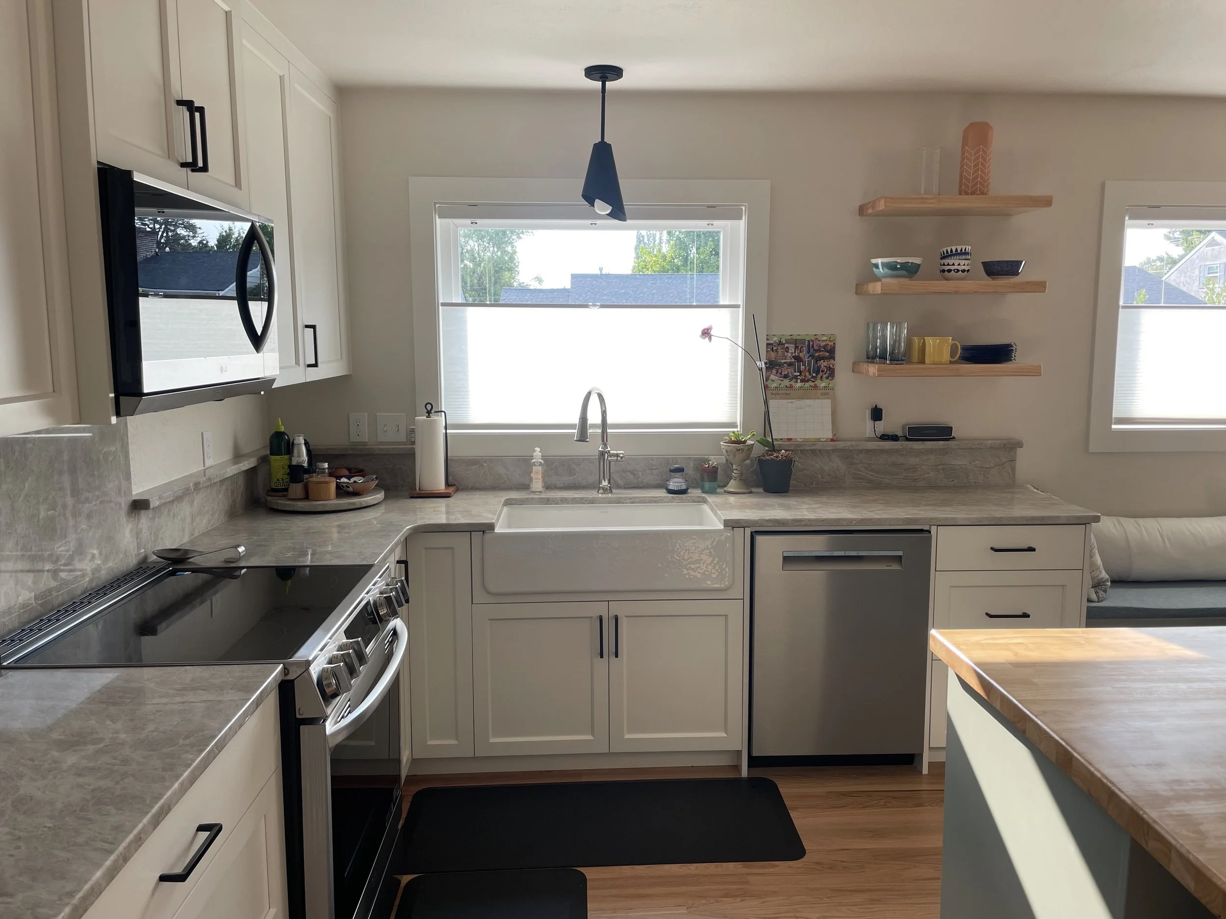 Modern kitchen with white cabinets, gray marble countertops, a window above the sink, and open wooden shelves with dishes, cups, and decorative items. There are appliances including a microwave, oven, and dishwasher, and a small seating area with a w