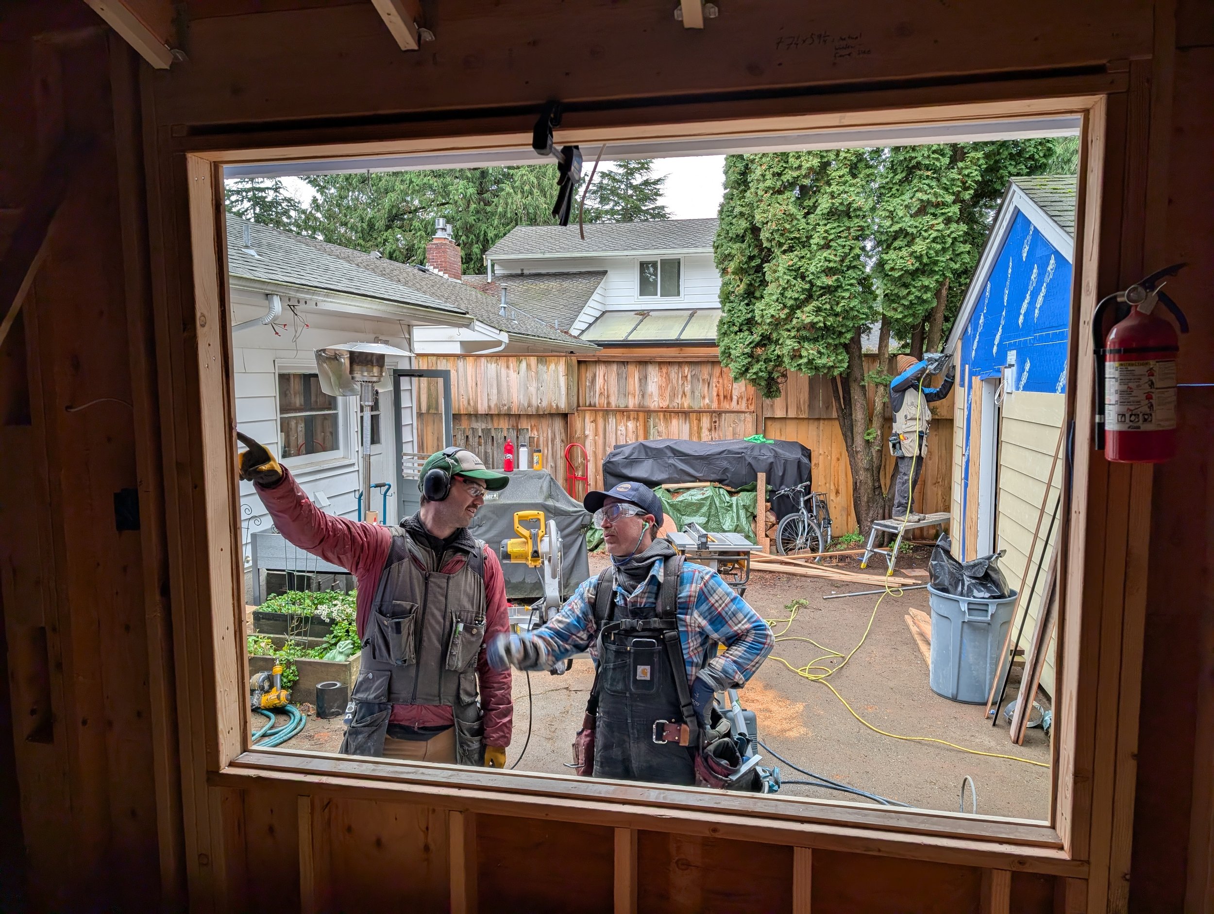 Two construction workers and a homeowner exchanging a fist bump outside a house under construction, viewed through a large window opening.