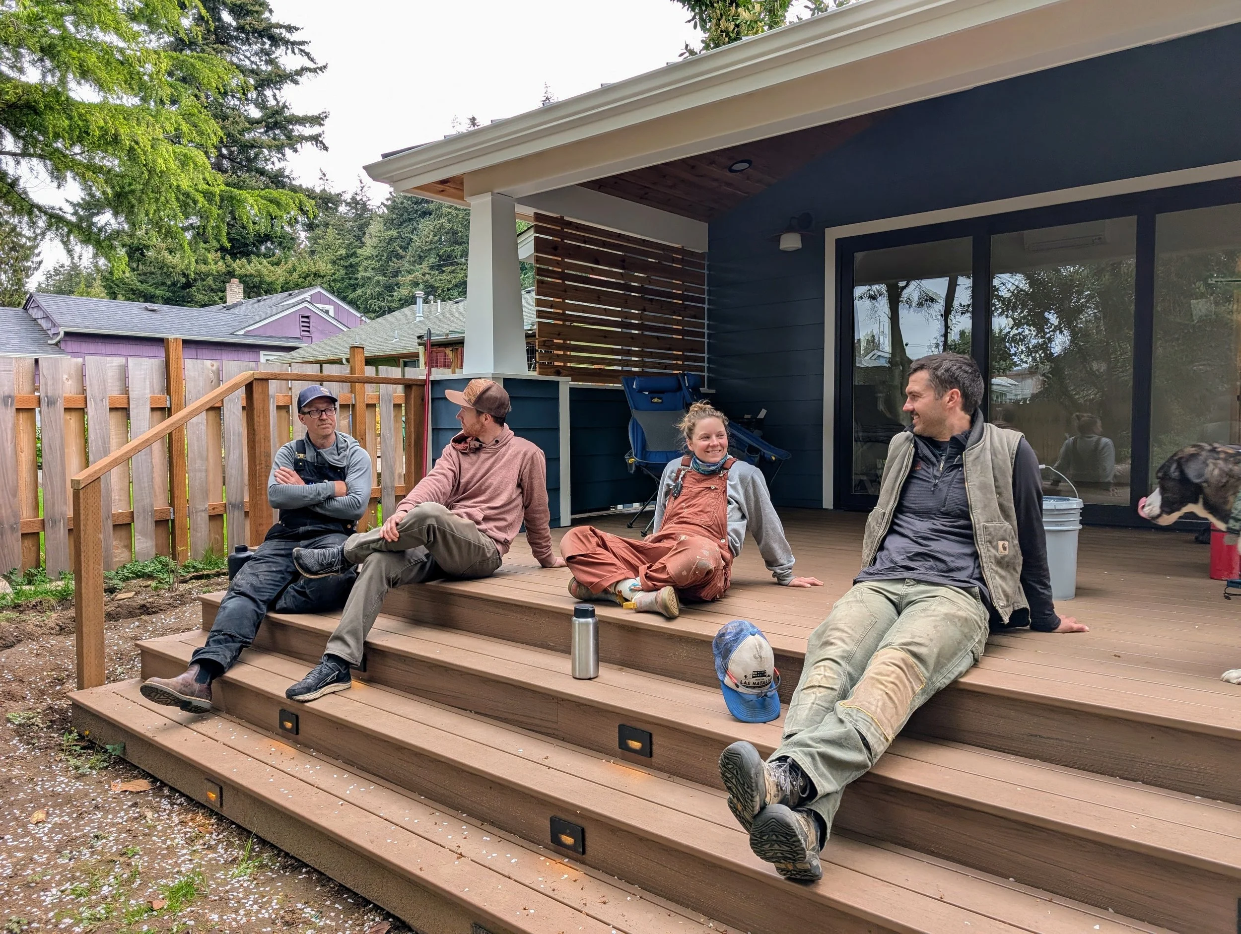 Four people sitting on a wooden deck outside a house, chatting and smiling, with some trees and neighboring houses in the background.