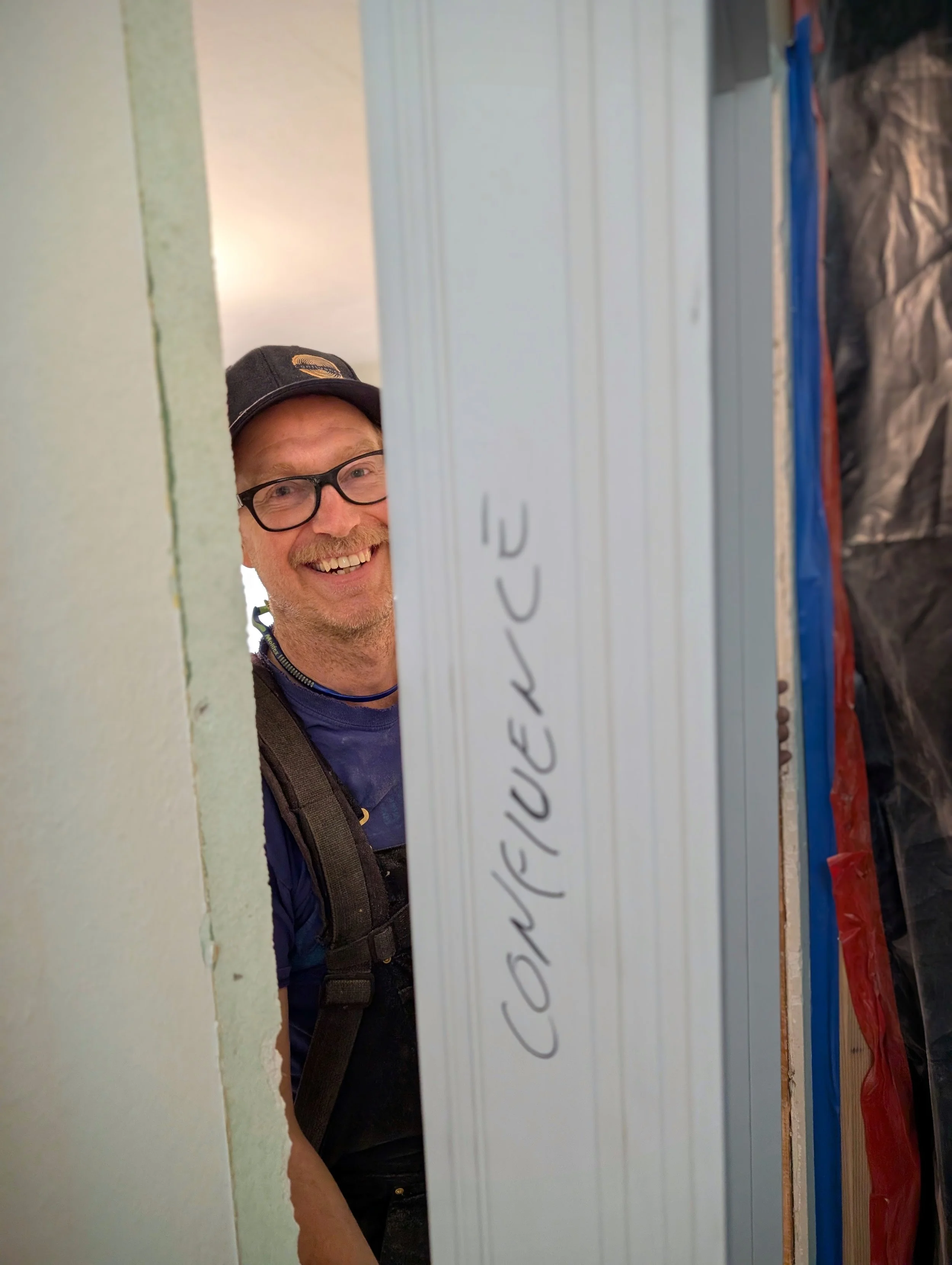Smiling man with glasses, beard, and cap looking through a hole in a wall during construction or renovation.