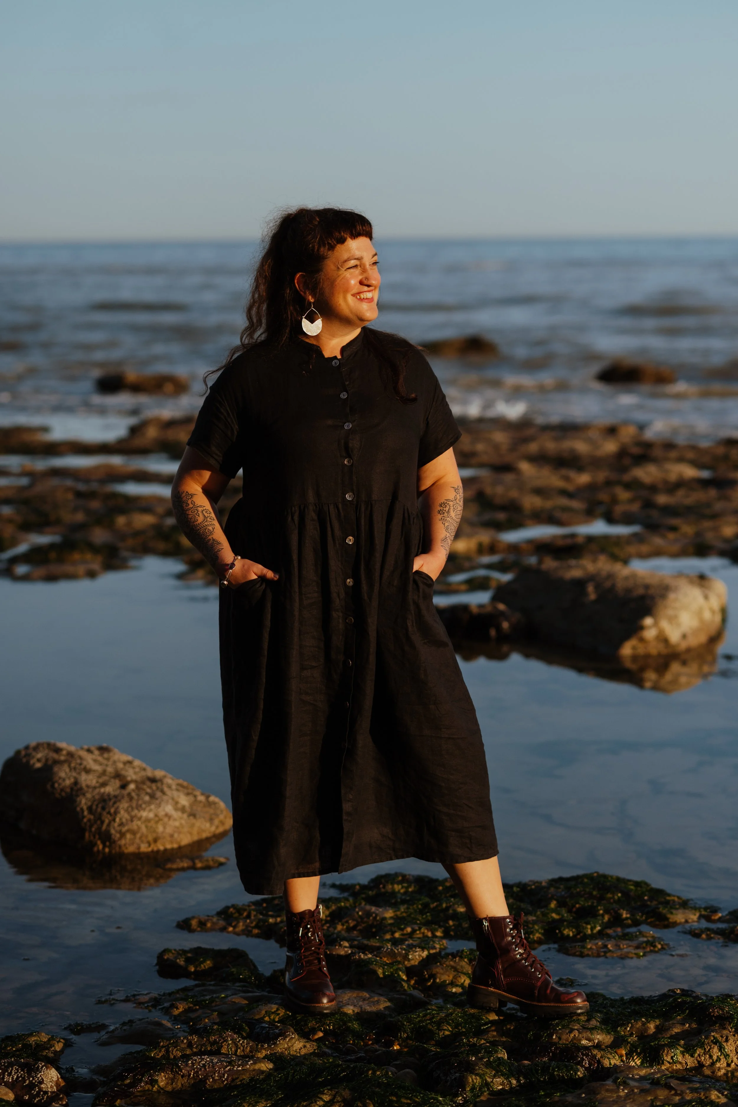 Woman standing on rocks by the ocean, smiling, with hands in pockets, wearing a black dress and brown boots.