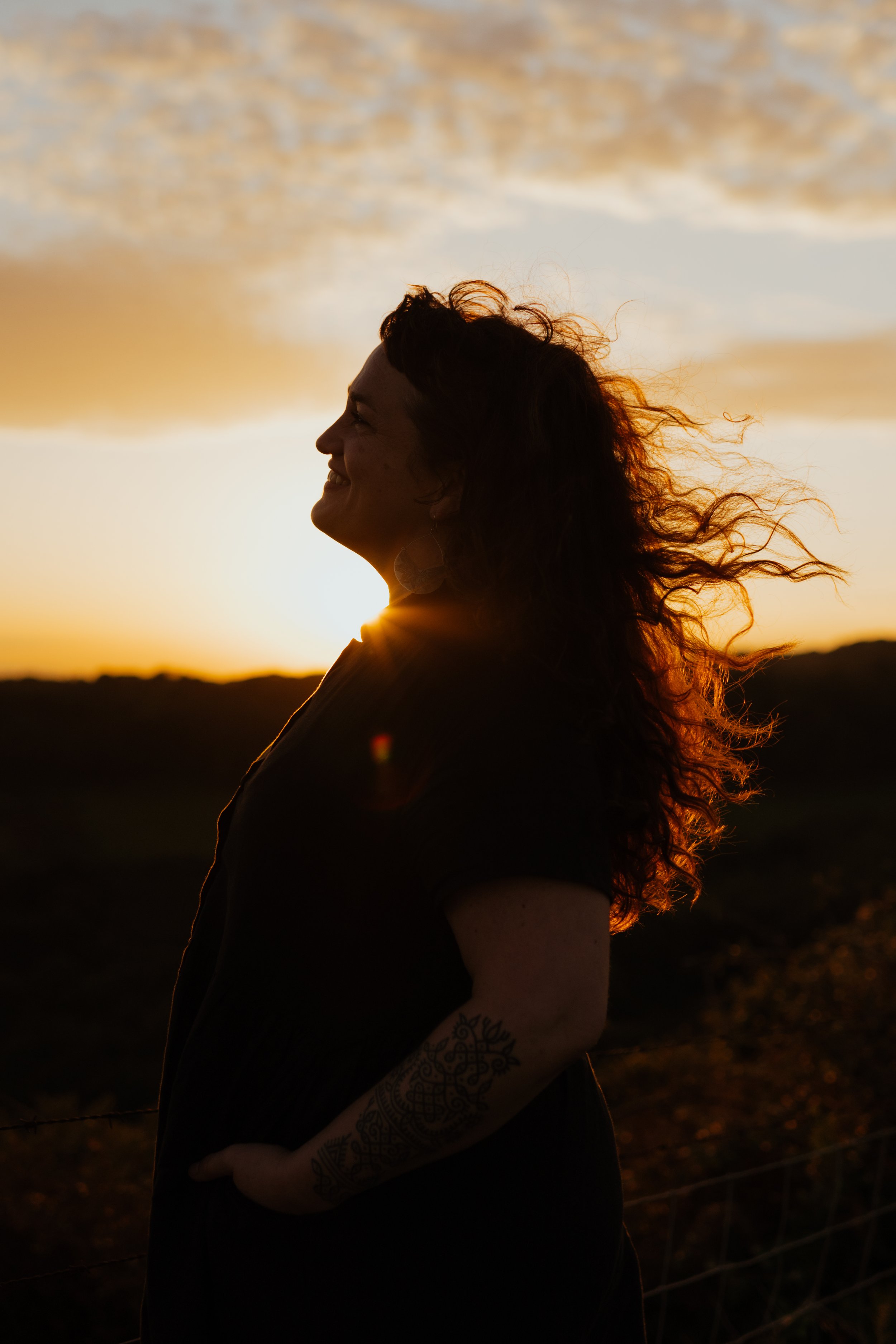 Silhouette of a smiling woman with curly hair standing outdoors at sunset.