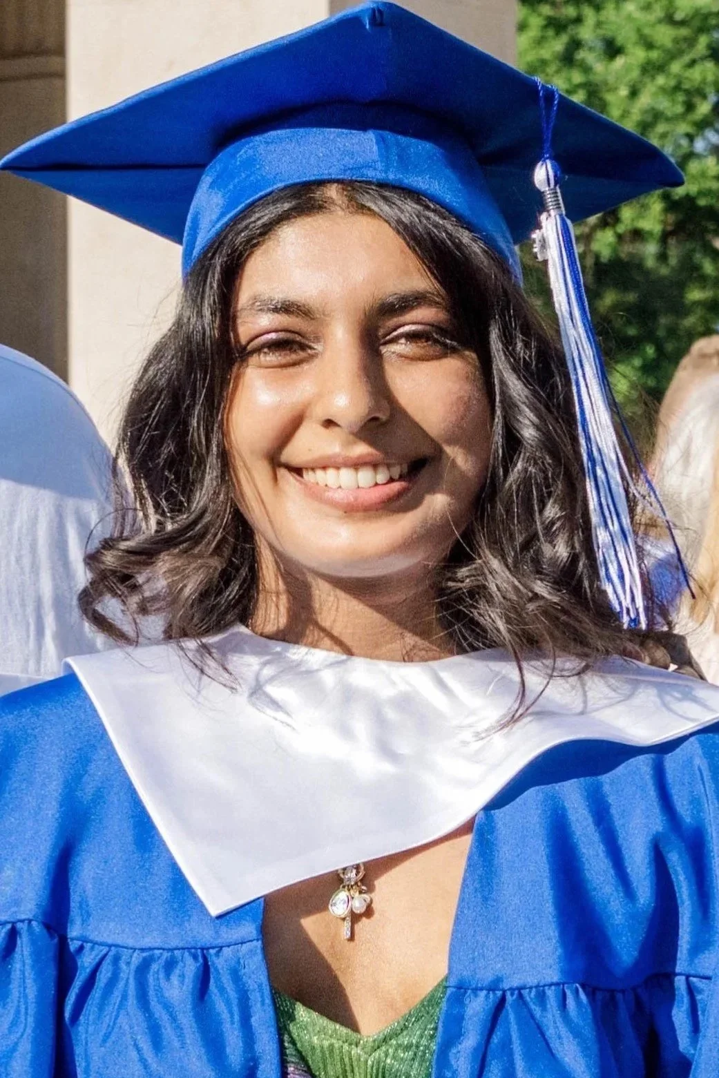 A young woman dressed in a royal blue graduation cap and gown, smiling outdoors with green trees in the background.