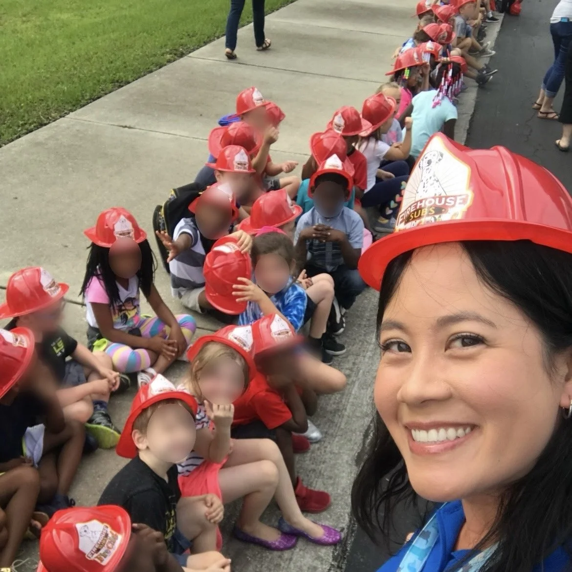 A group of children sitting on the sidewalk wearing red firefighter helmets, with Ms. Lo in the foreground smiling for a selfie, also wearing a firefighter helmet, during a fire station visit or safety event.