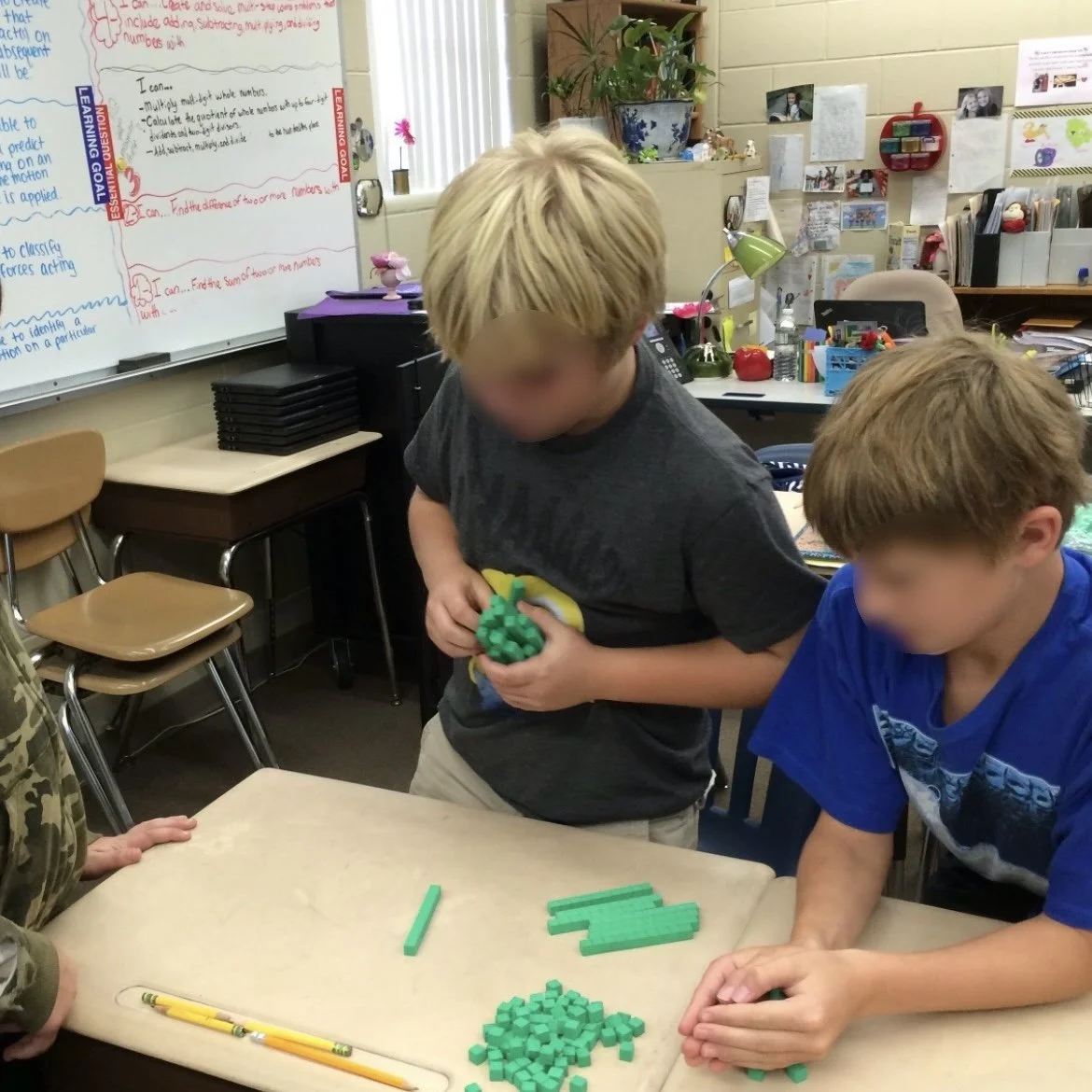 Ms. Lo's former 5th grade students during her math manipulative activity: Two young boys working together at a classroom table, handling green math manipulatives, with a whiteboard in the background containing handwritten notes and learning goals.