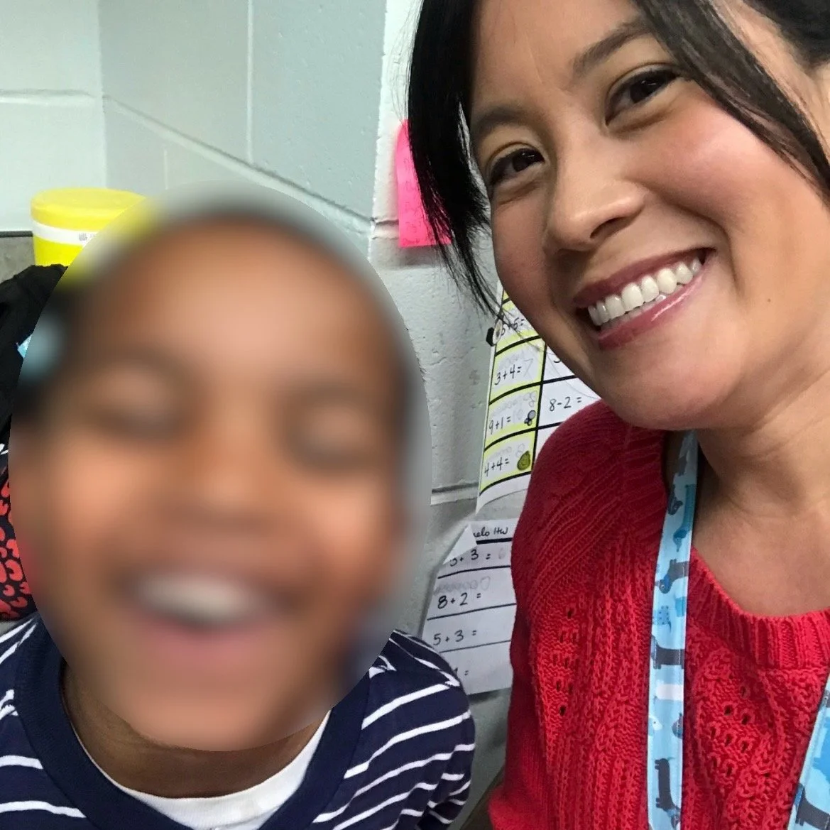 Ms. Lo with a former student: A smiling woman with black hair and a red sweater taking a selfie with a child whose face is blurred, in a classroom setting with a whiteboard in the background.