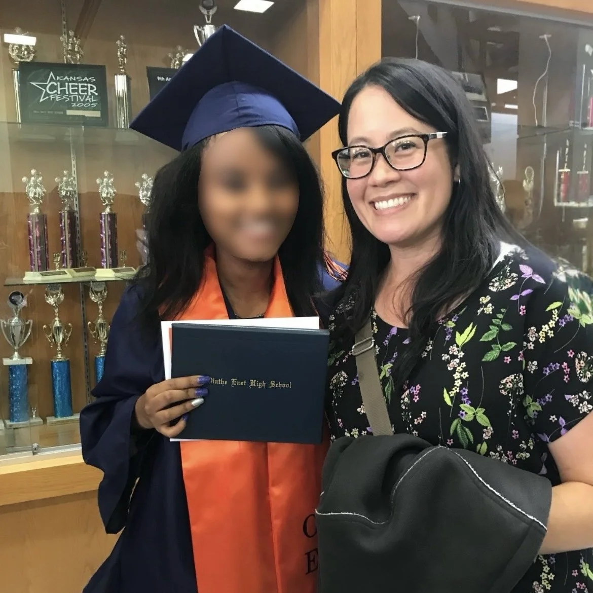 Ms. Lo and her former student at the student's high school graduation: A young woman in a graduation gown and cap holding a diploma, smiling, standing next to an adult woman in a floral shirt, also smiling, beside a trophy display case.