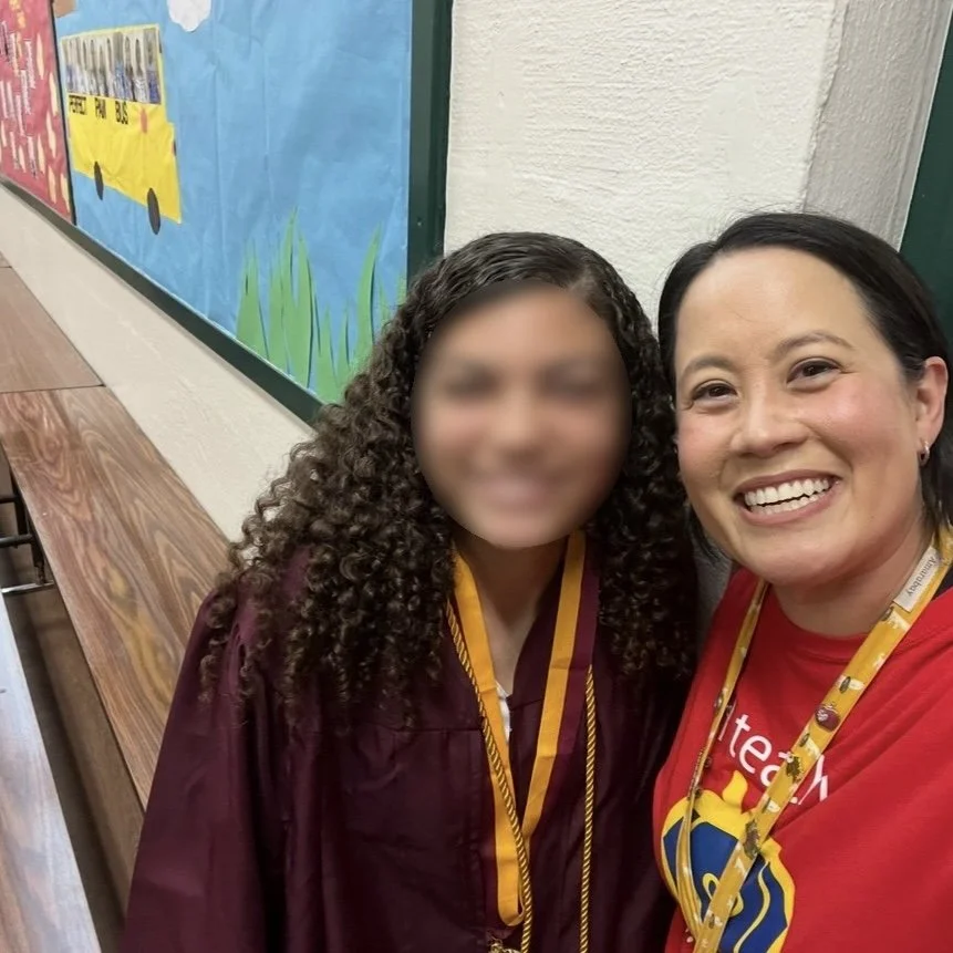 Ms. Lo and her former student in her high school graduation regalia: A high school graduate with her elemetary teacher smiling for a selfie in a school hallway, with colorful bulletin boards and wooden benches in the background.