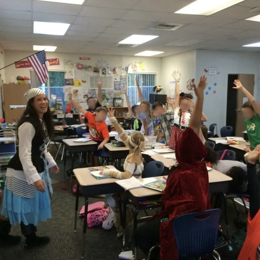 Ms. Lo and former 5th grade class on Halloween: Classroom of elementary school students raising hands while teachers talk, decorated with American flags and colorful posters.